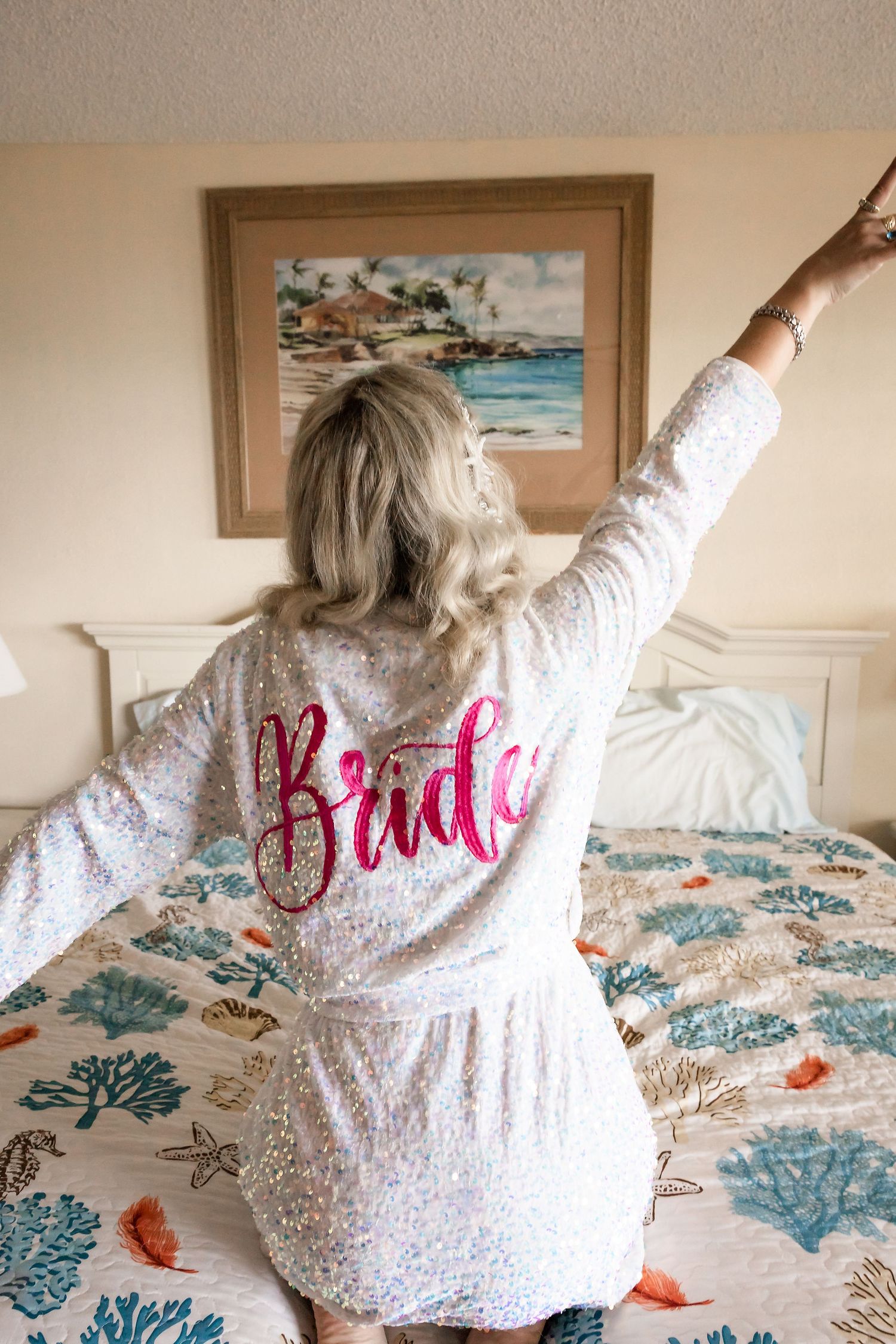 Blonde bride in a sparkly white Bride robe stretching arms up on a bed in a beach hotel room.
