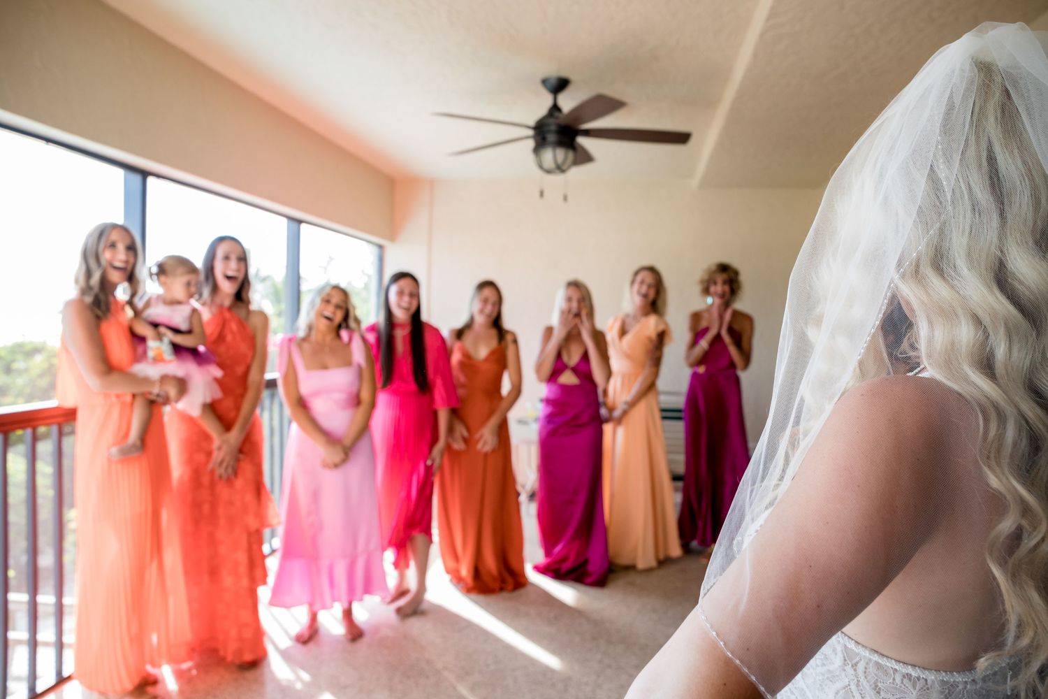 Bride in white gown and veil reveals herself to bridesmaids in colorful coral and pink dresses.