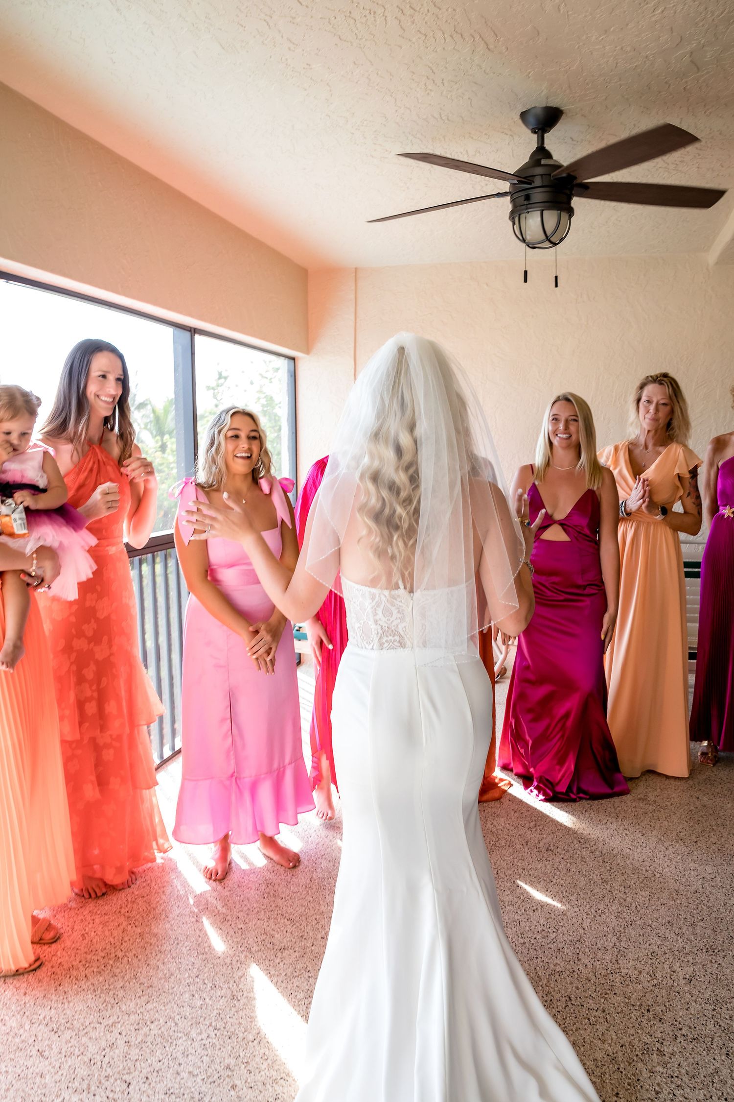 Bride in white fitted gown and veil surrounded by bridesmaids in vibrant pink and orange dresses.
