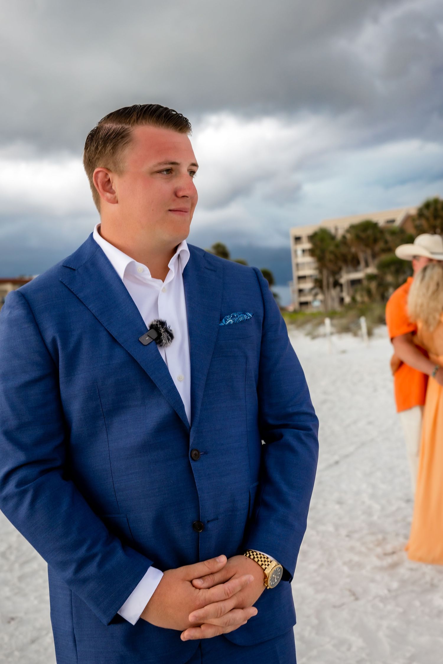 Groom in blue suit standing on white sand beach before wedding ceremony under cloudy skies.