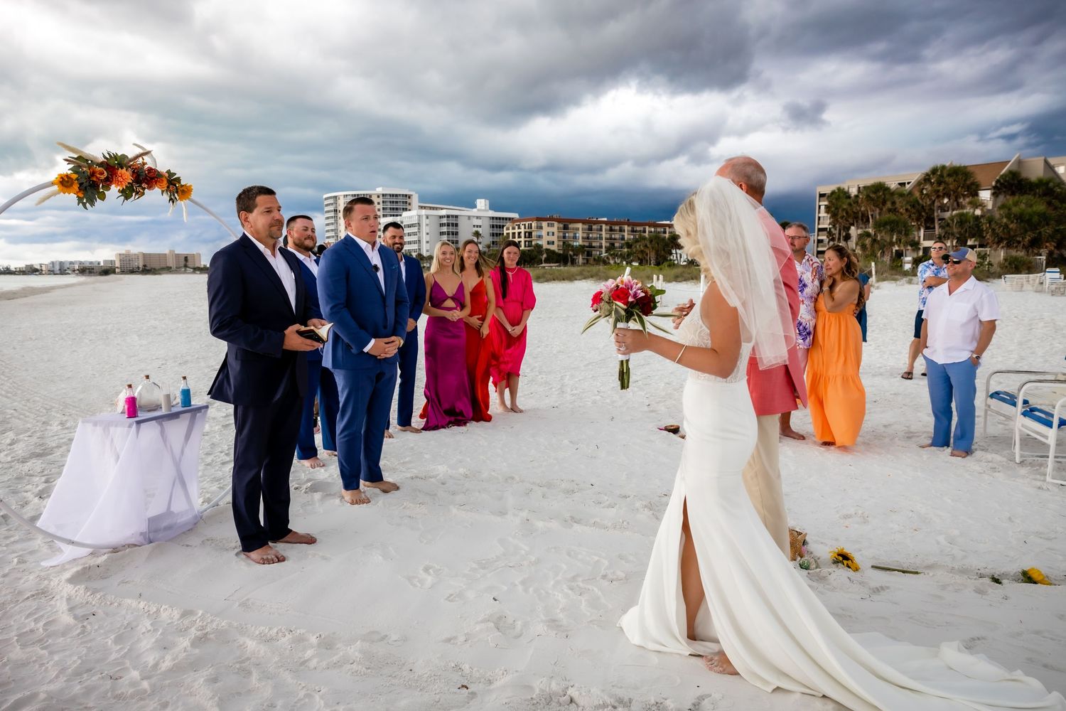 Bride walks toward groom and wedding party during outdoor beach ceremony under dramatic clouds.