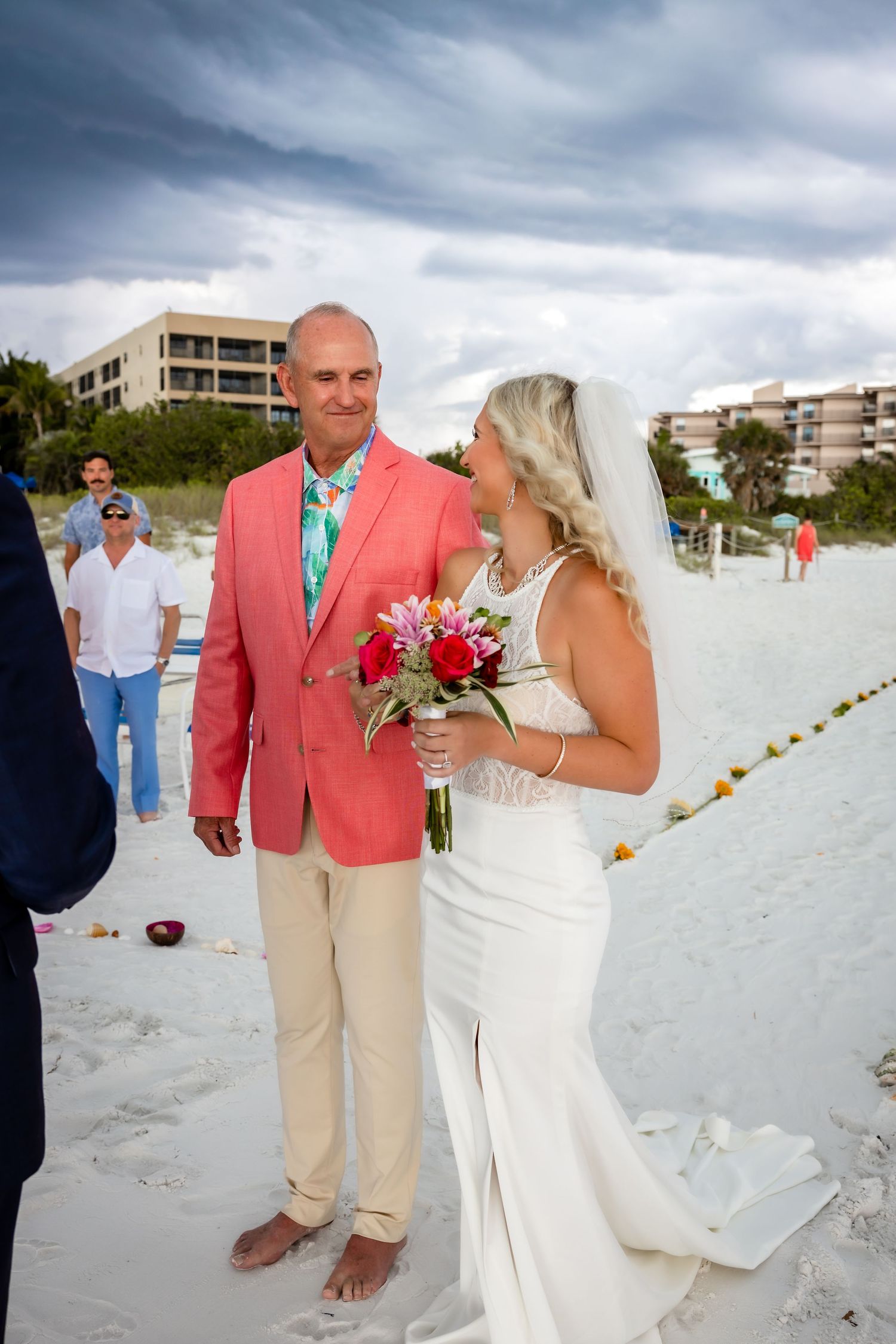 Father escorts smiling bride holding colorful bouquet across white sand beach at wedding.