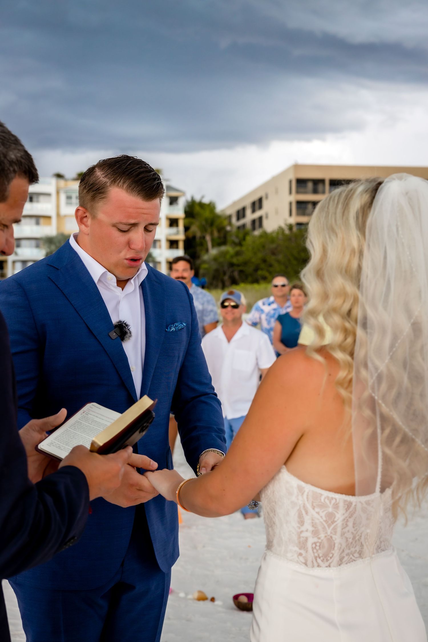 Groom in blue suit recites vows from book to bride during emotional beach wedding ceremony.