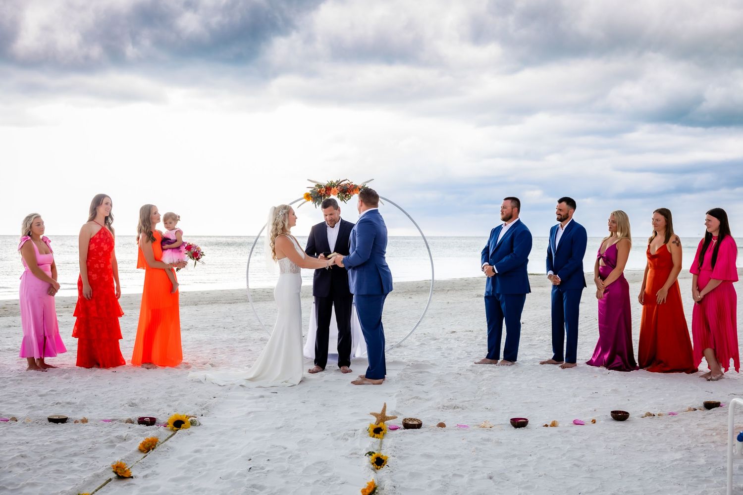 Bride and groom exchange vows under floral arch flanked by colorful bridesmaids and groomsmen.