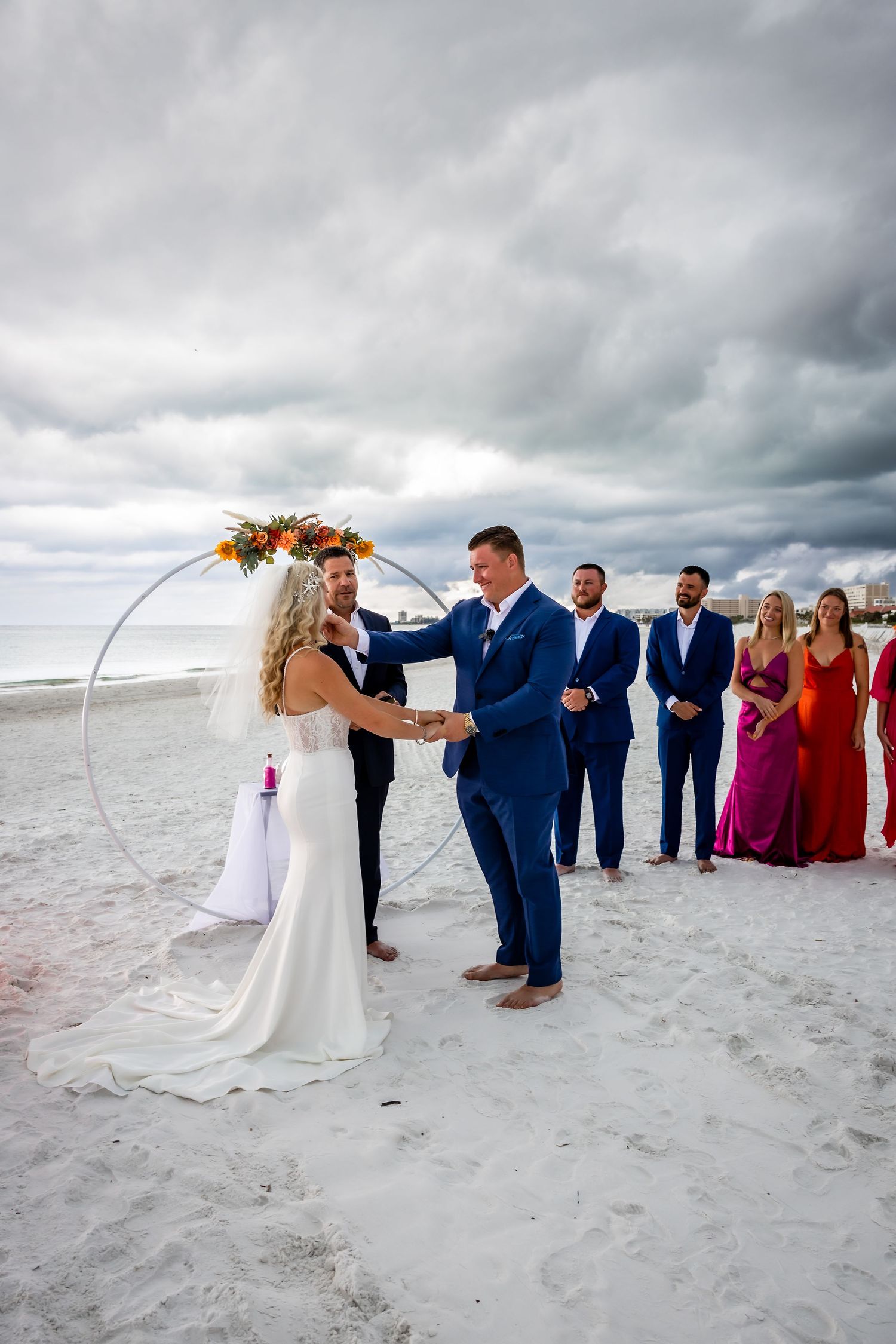Couple holds hands at circular floral arch during beach wedding with bridal party looking on.