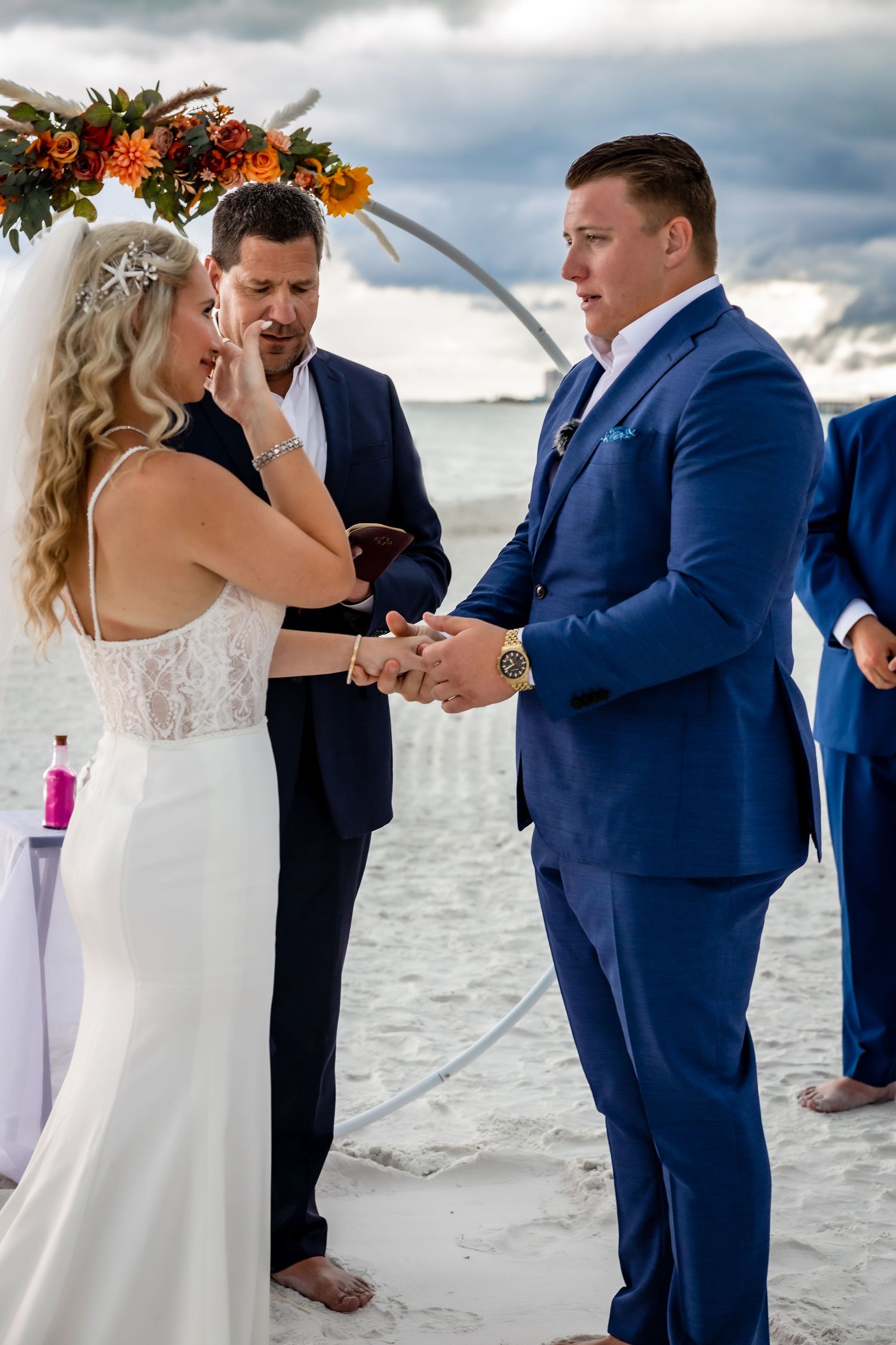 Emotional bride wipes tear while holding grooms hands during heartfelt beach wedding ceremony.