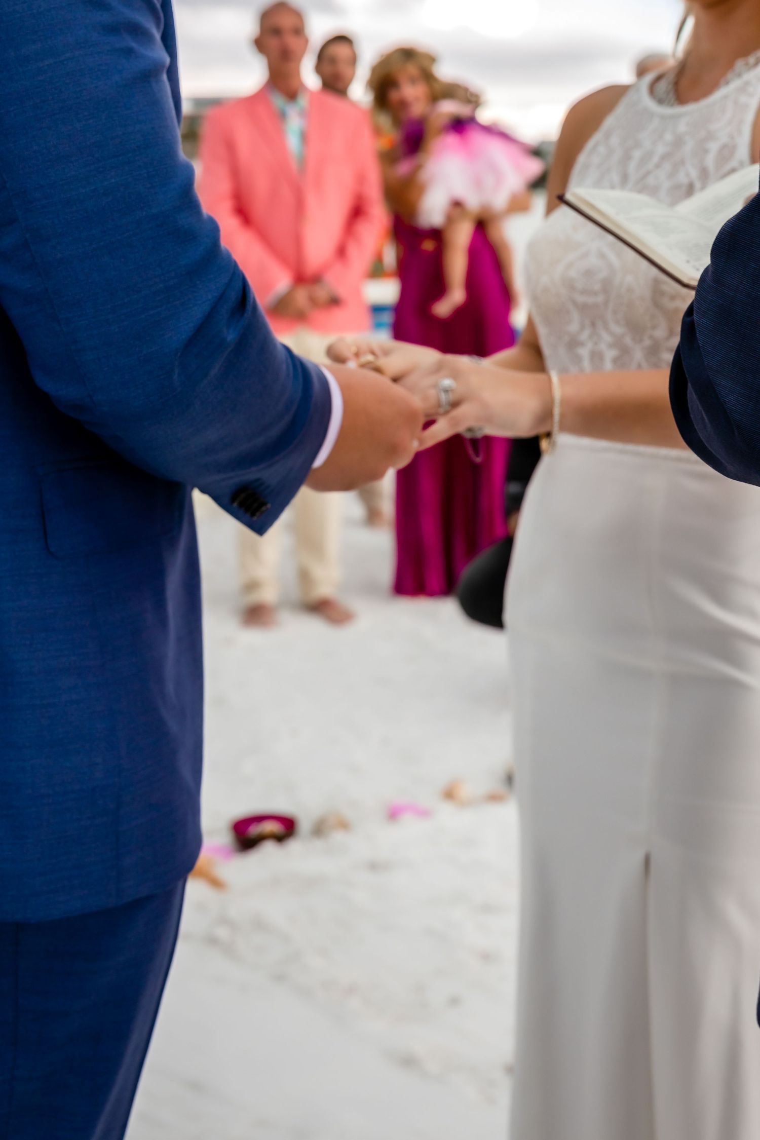 Close-up of bride and groom holding hands during ring exchange at beach wedding ceremony.