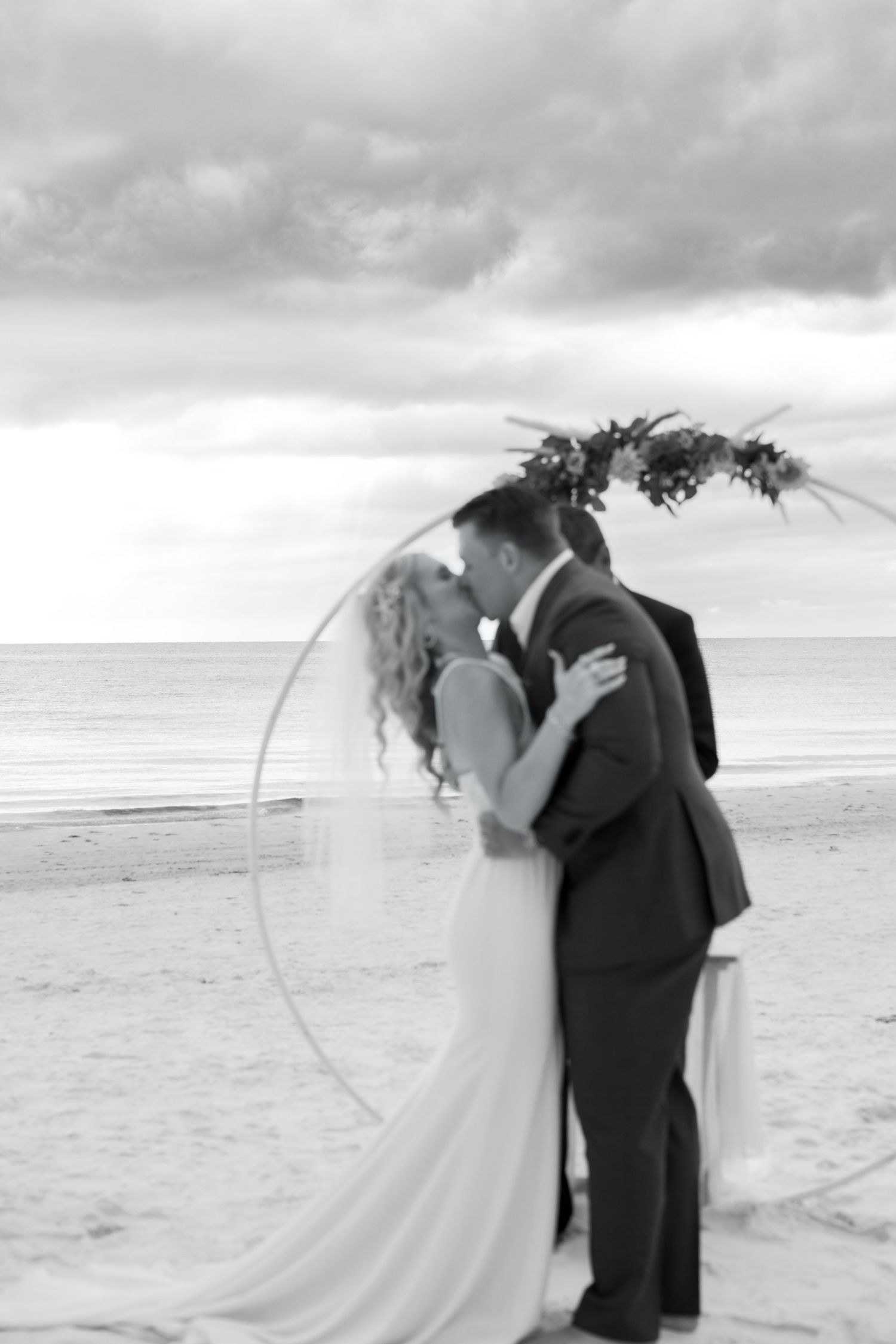 Black and white photo of newlyweds sharing first kiss under floral arch on the beach.