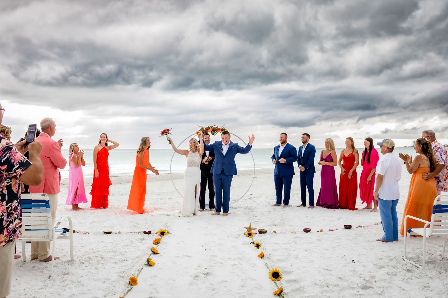 Bride and groom celebrate at beach wedding ceremony with colorful wedding party on white sand under dramatic cloudy skies.