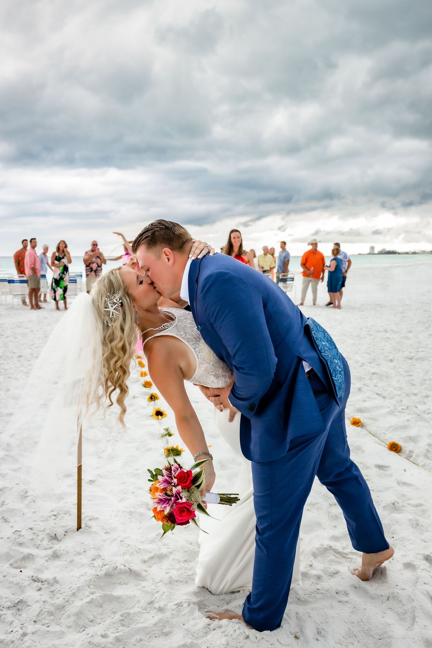Groom dips bride for romantic kiss on white sand beach after outdoor wedding ceremony with sunflower aisle decor.