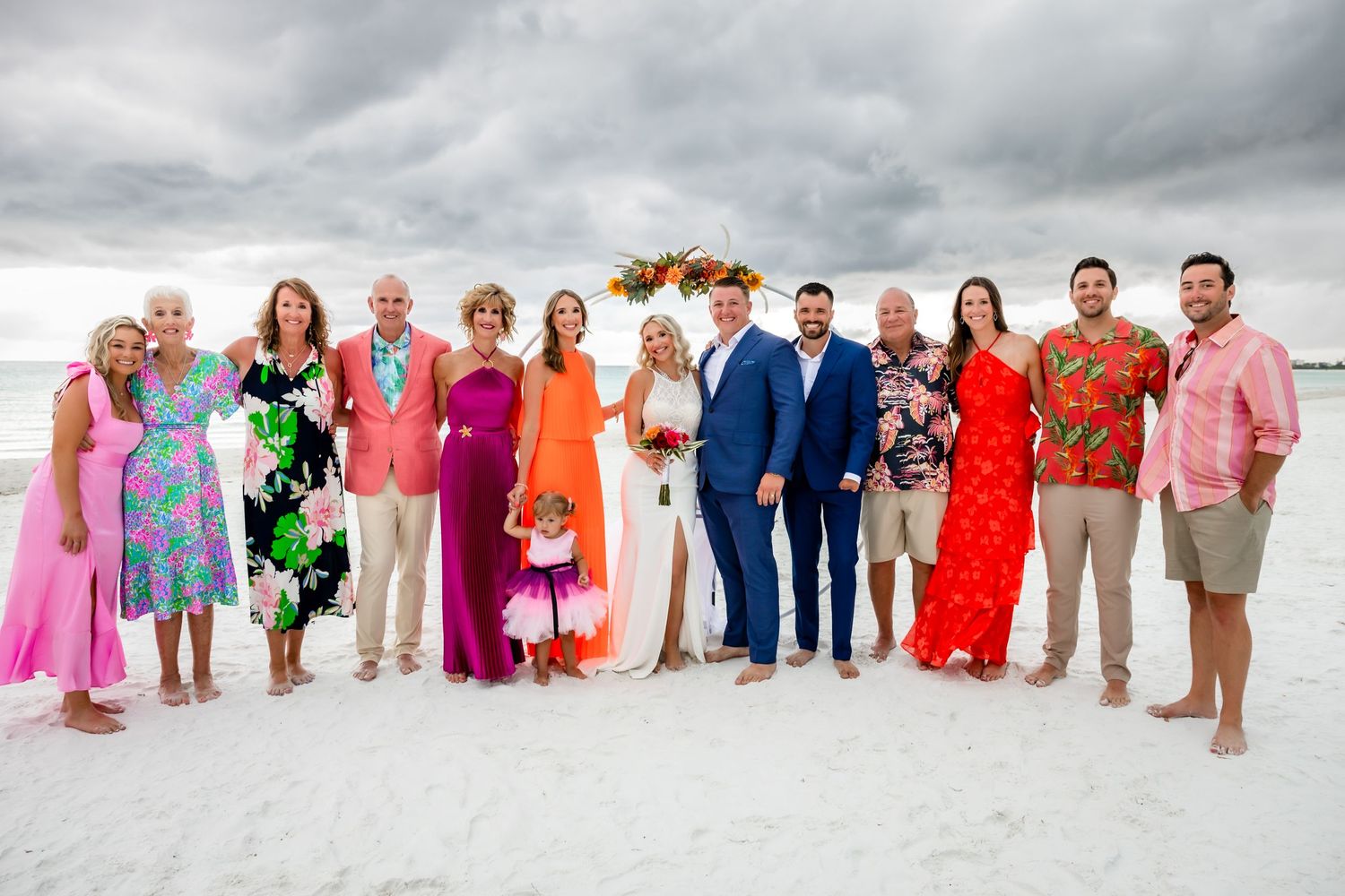 Newlyweds pose with family group portrait on white sand beach after colorful destination wedding ceremony.
