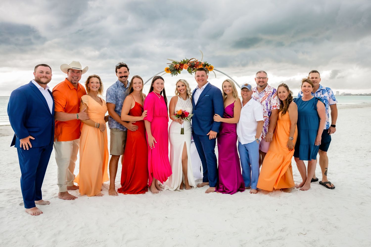 Wedding party and family pose together on white sand beach in vibrant colorful attire after beach ceremony.