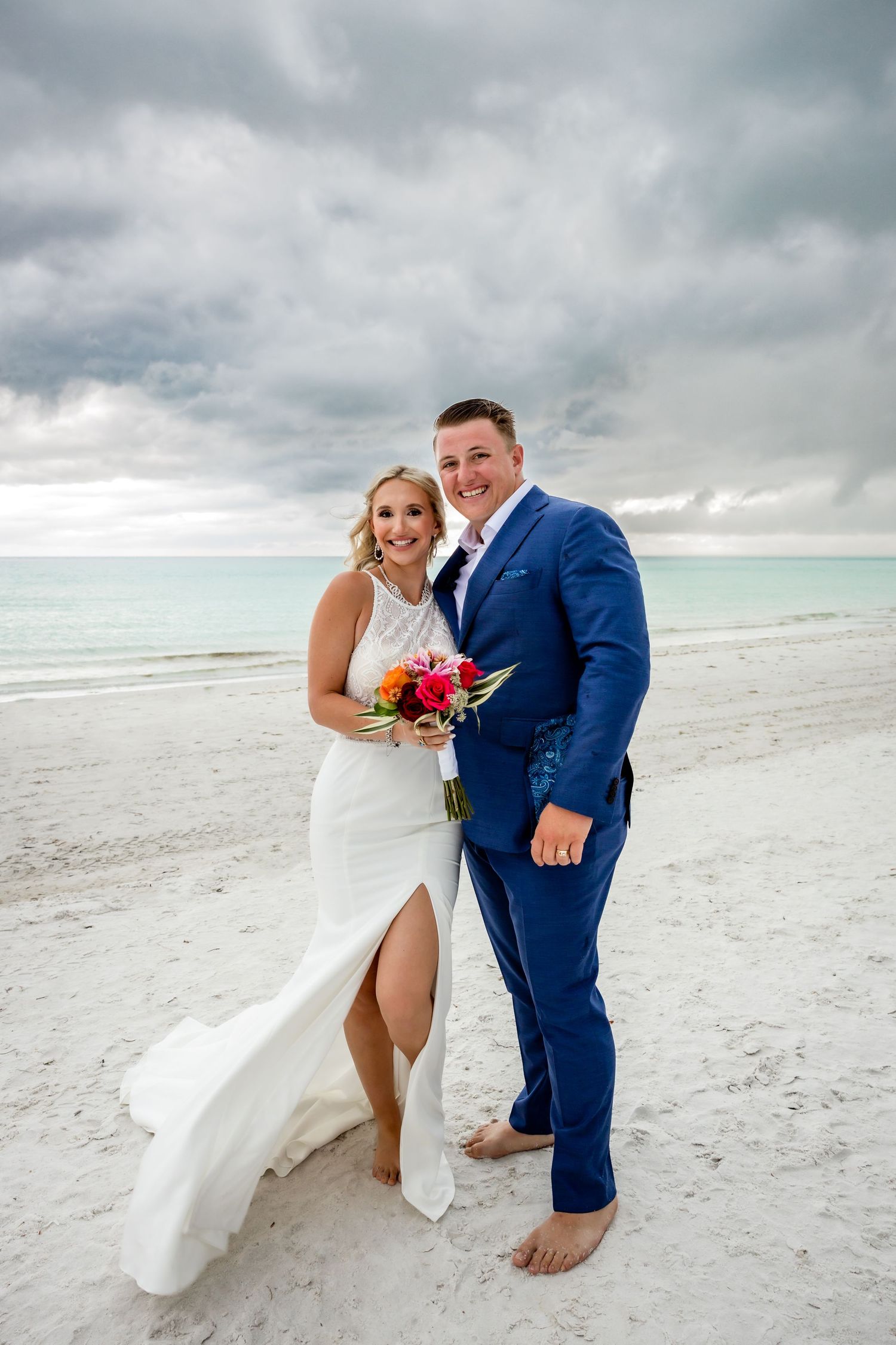 Smiling bride holding colorful bouquet and groom in blue suit pose together on white sand beach after wedding.