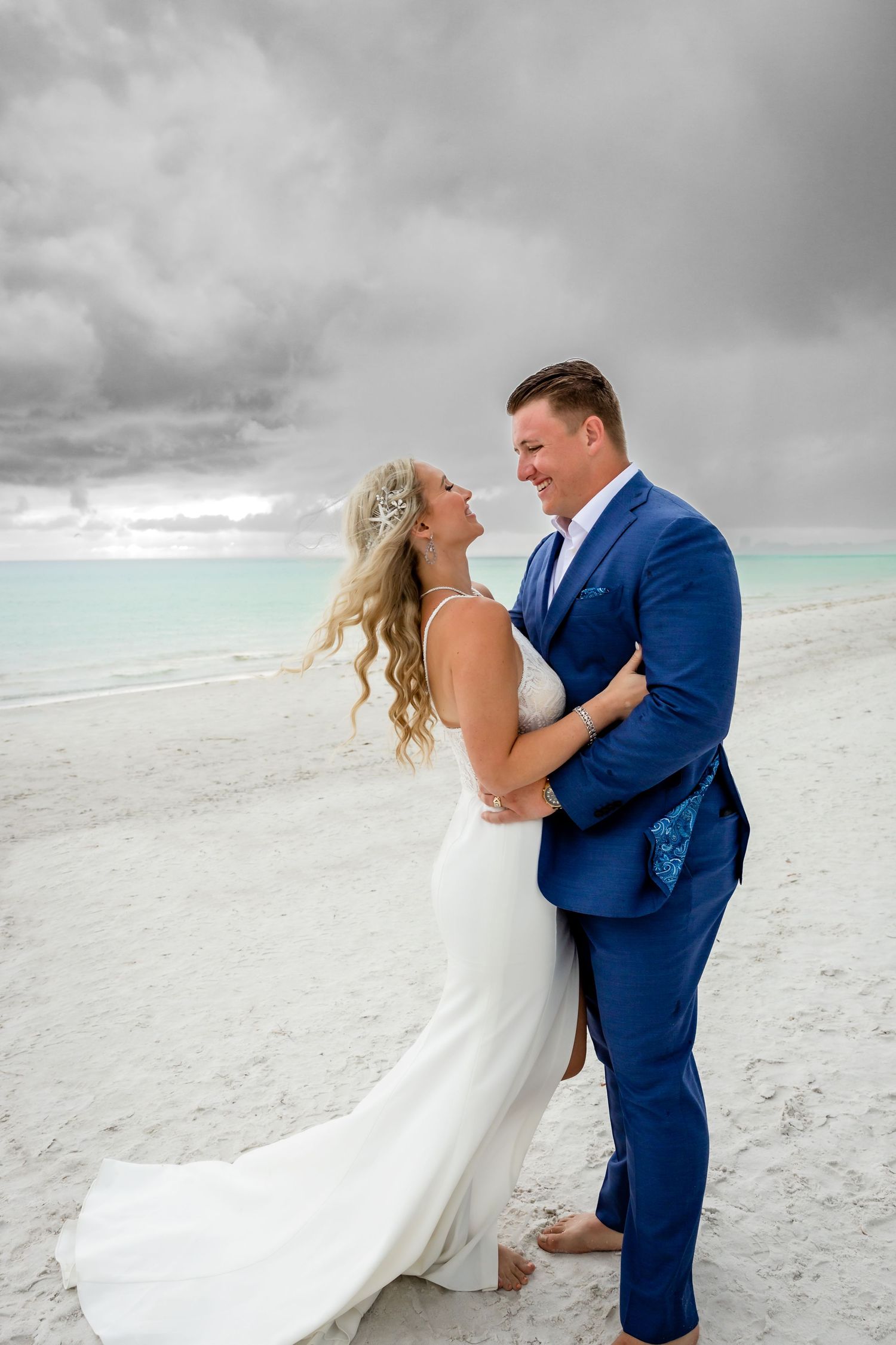 Bride in white gown and groom in blue suit share romantic moment on white sand beach under stormy skies.