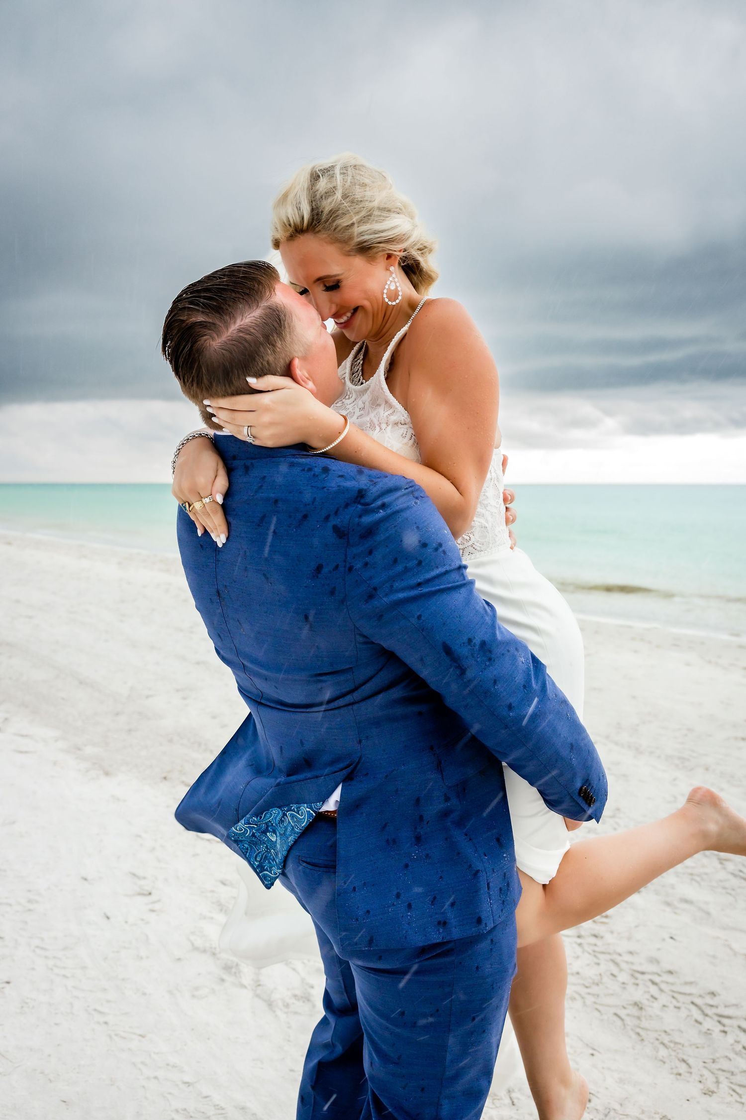 Groom in blue suit lifts laughing bride on white sand beach with turquoise ocean in the background.