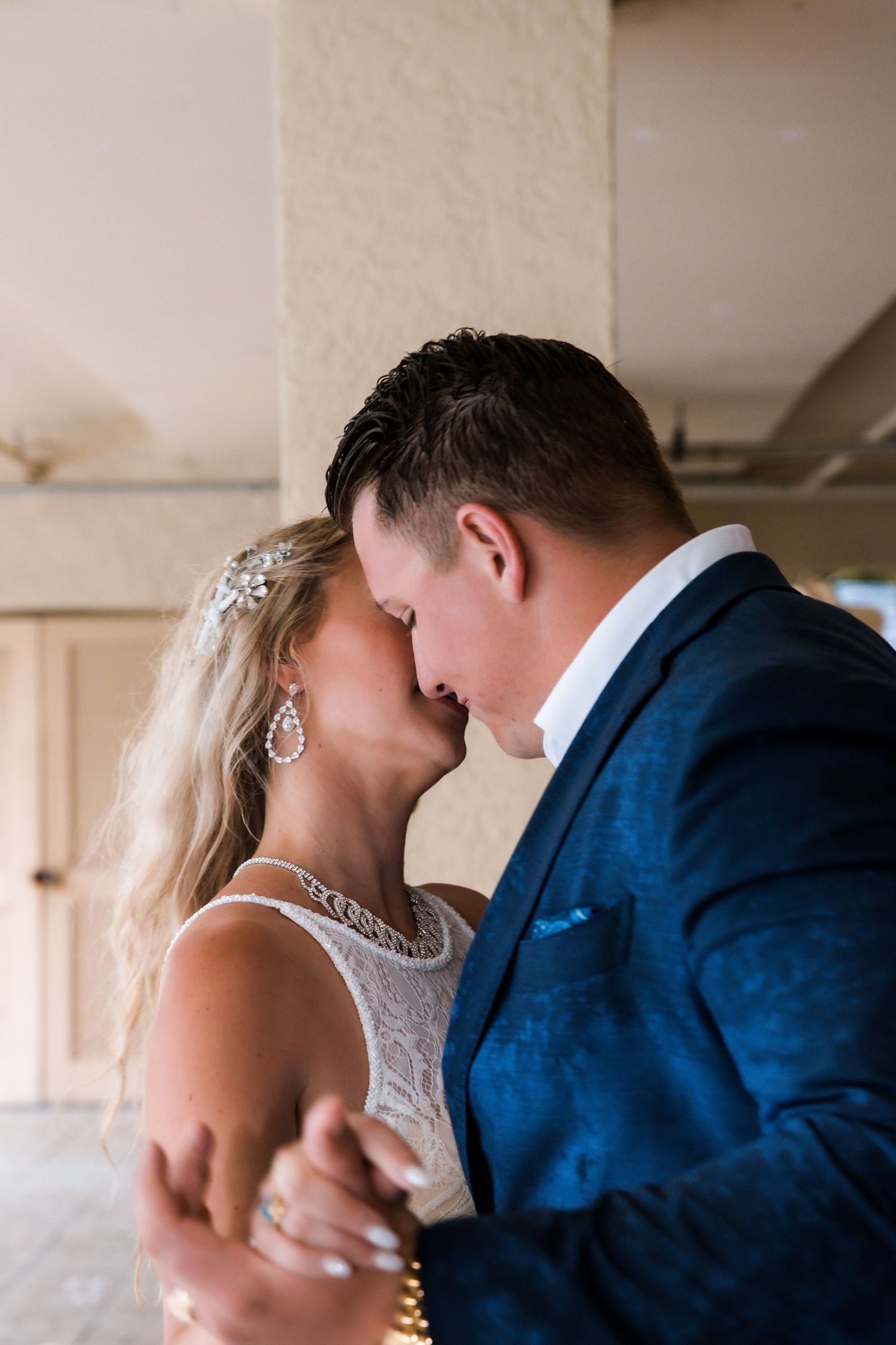Bride wearing crystal hair piece and earrings shares tender moment with groom in blue suit during first dance.