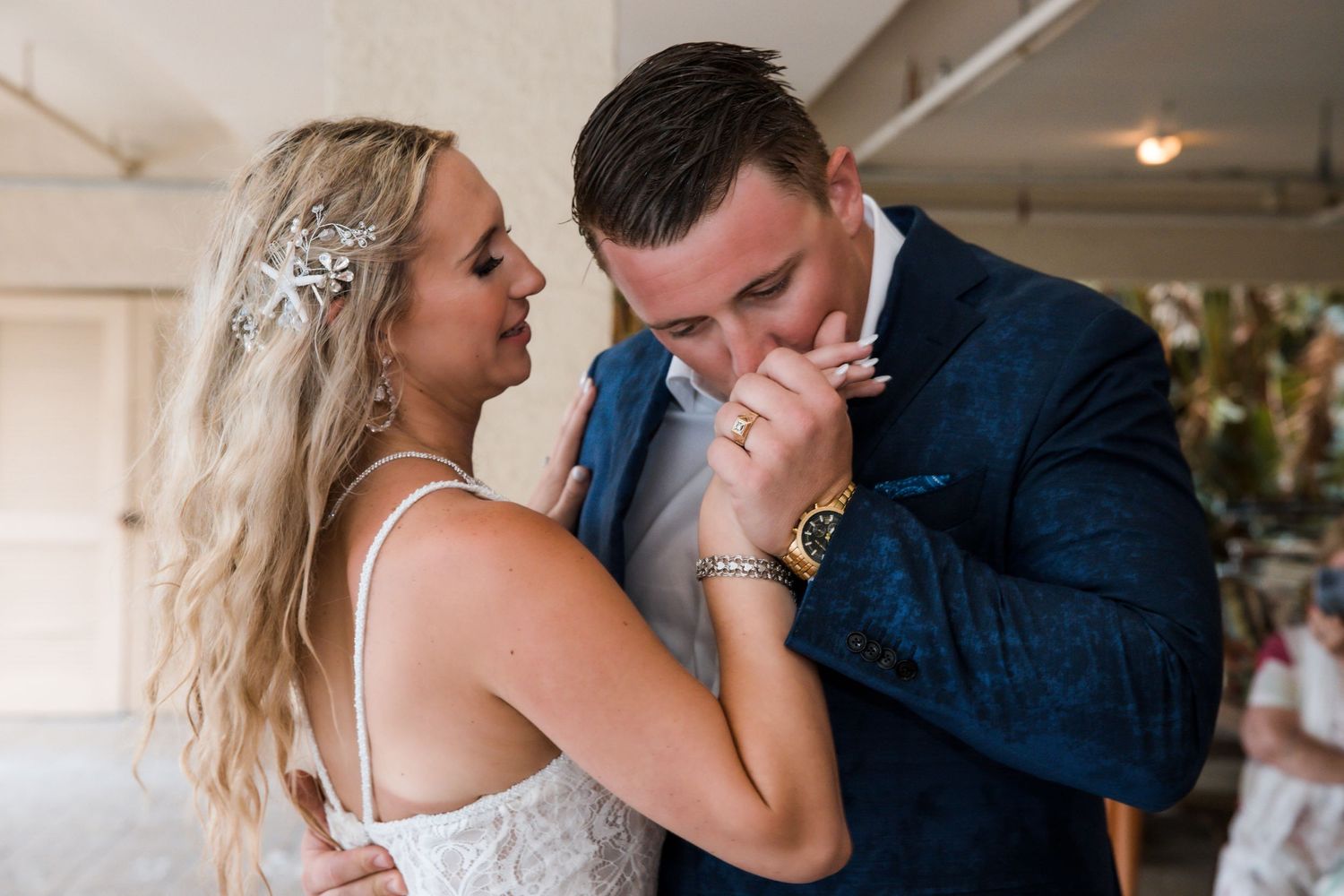 Emotional groom in blue suit kisses brides hand during wedding reception as she tenderly touches his face.