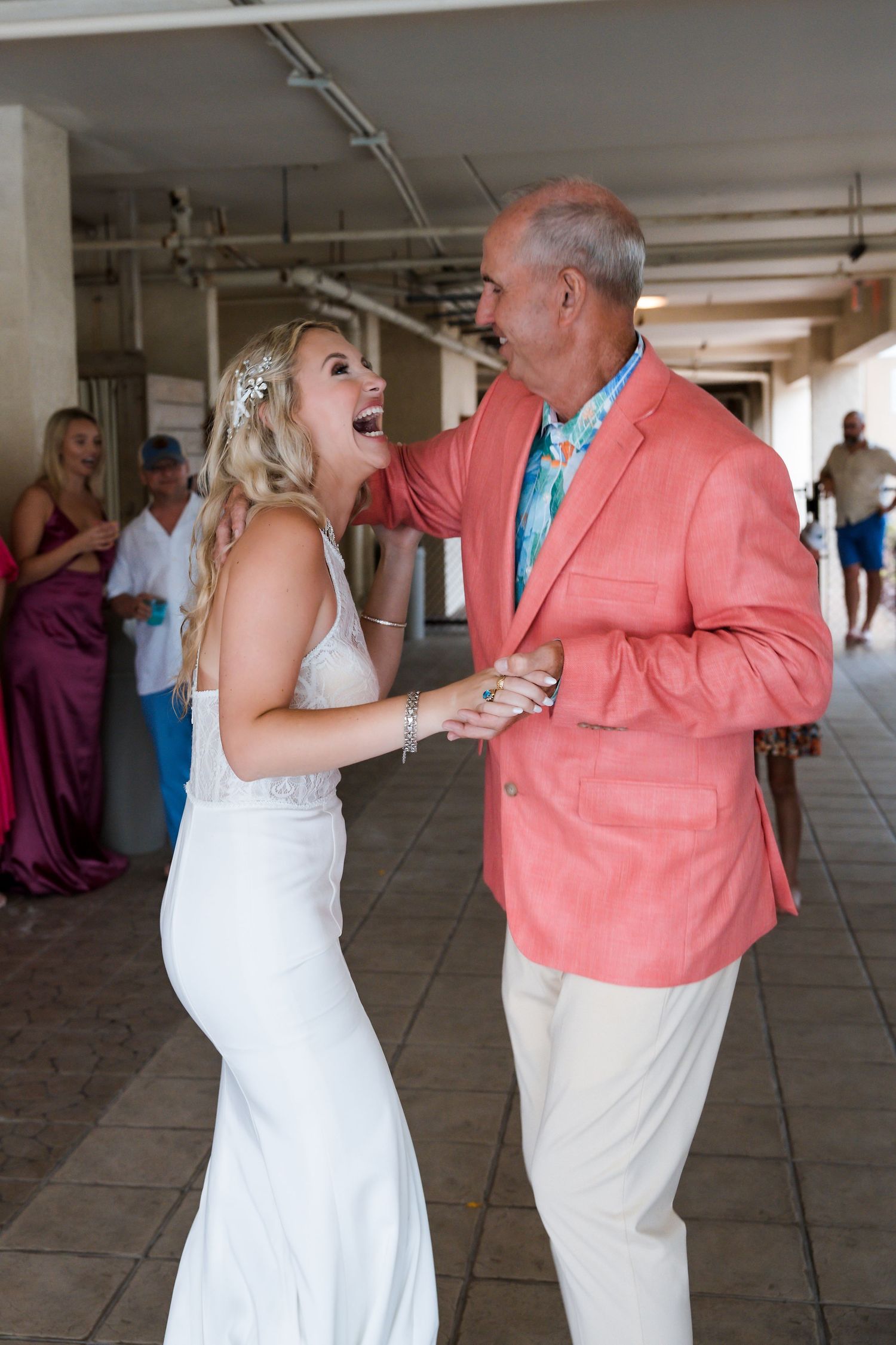Bride in white gown laughs joyfully with father in coral blazer during wedding dance.