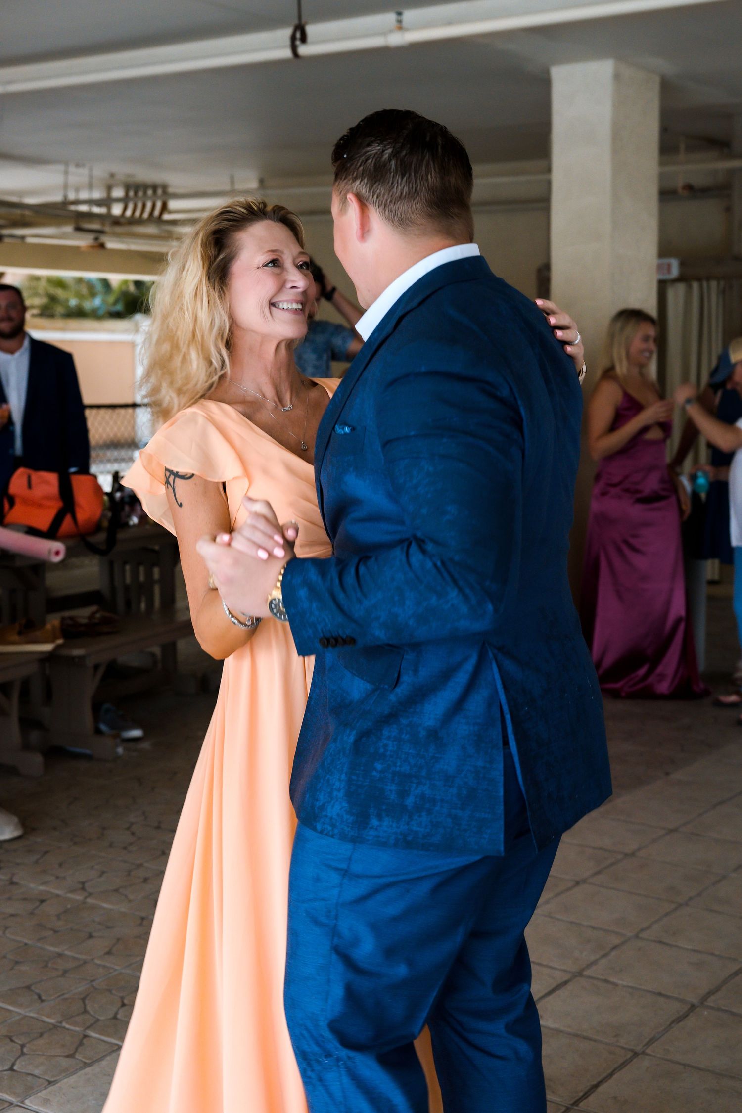 Groom in blue suit shares mother-son dance with woman in peach gown at wedding.