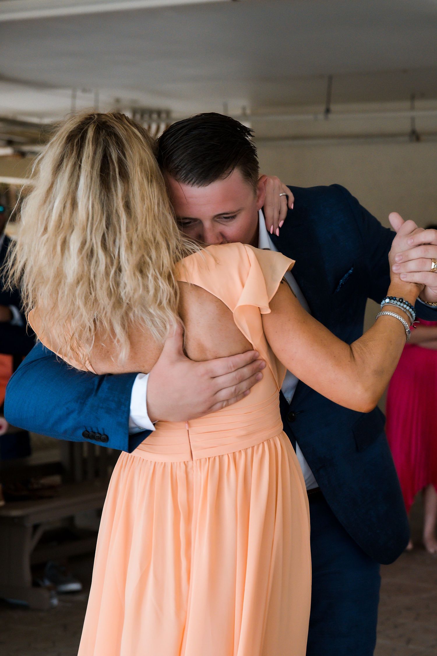 Groom in navy suit embraces mother in peach dress during emotional wedding reception dance.