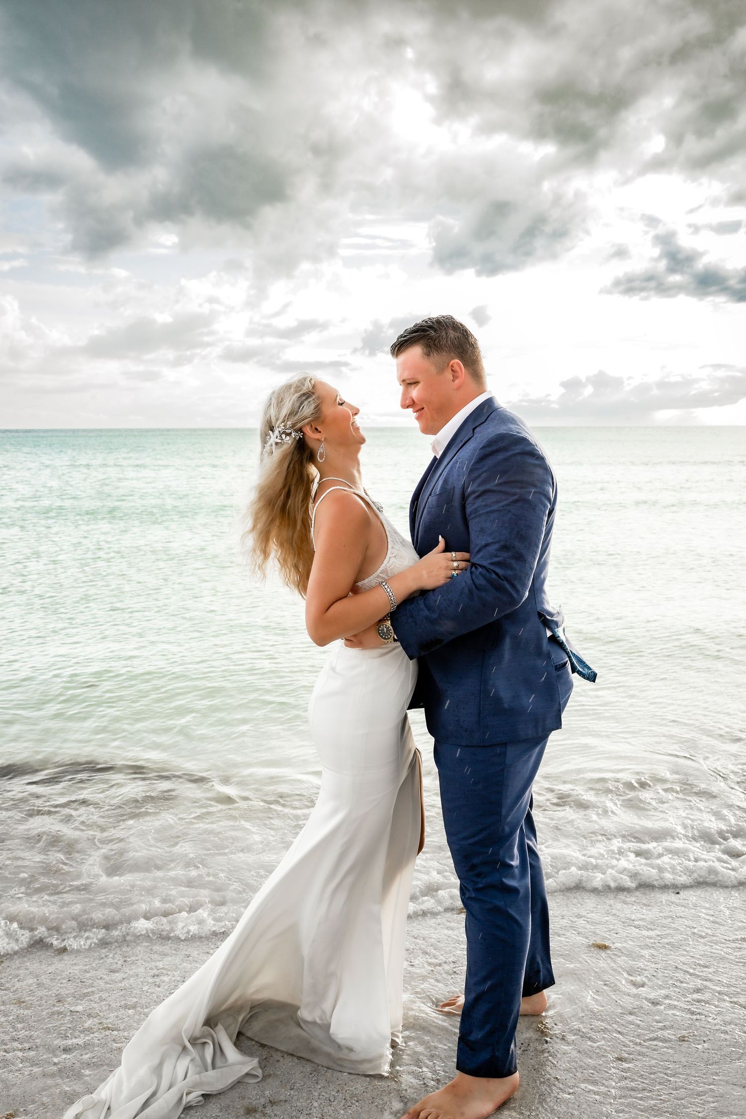 Bride and groom embrace barefoot on beach with dramatic cloudy sky and ocean backdrop.