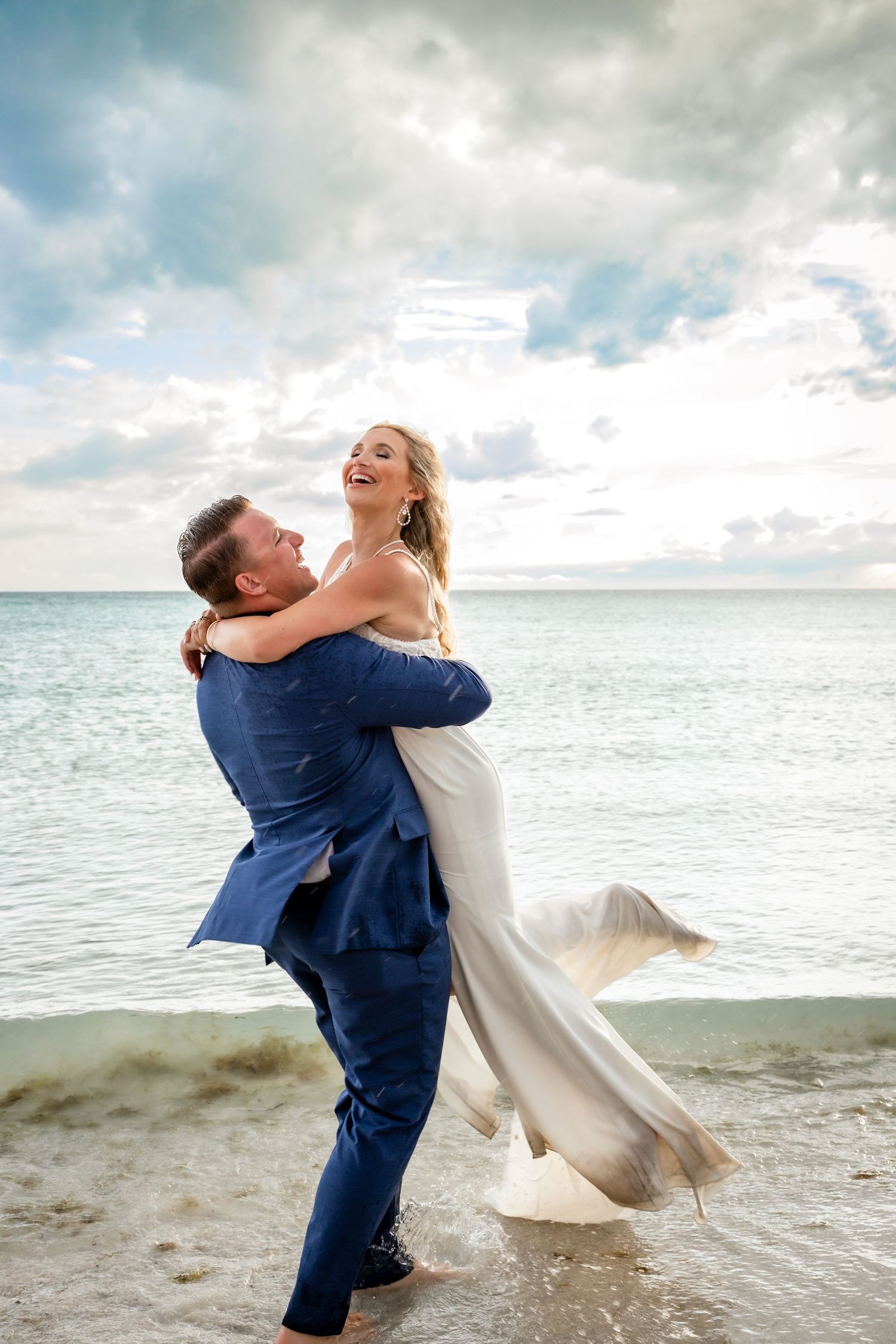 Groom lifts laughing bride in white gown on beach with ocean and cloudy sky behind.