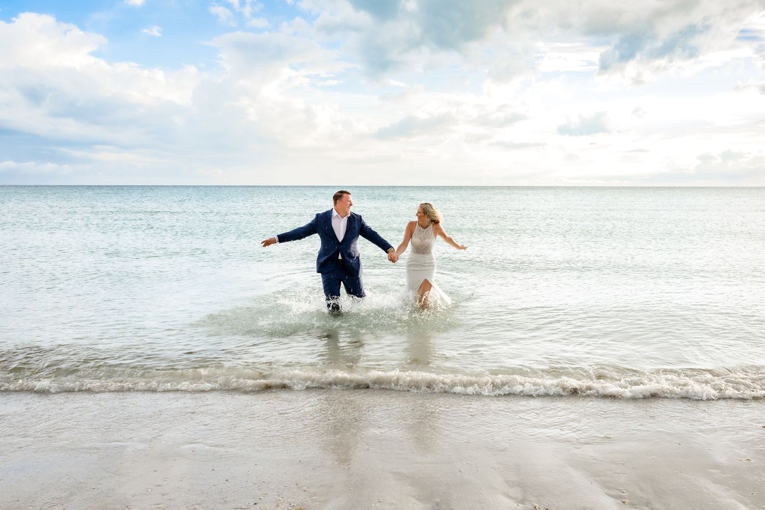 Bride and groom hold hands while splashing through ocean waves on beach after wedding.