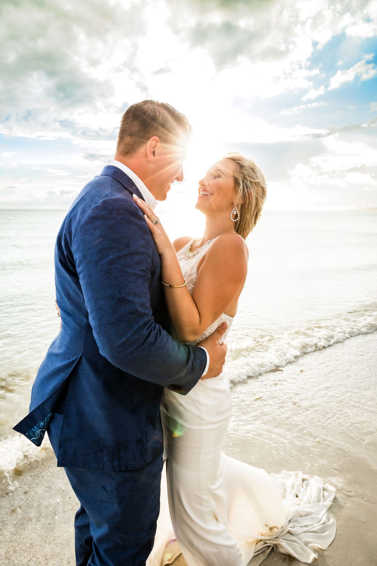 Bride and groom embracing on a sunny beach with ocean waves, sharing a joyful moment on their wedding day.