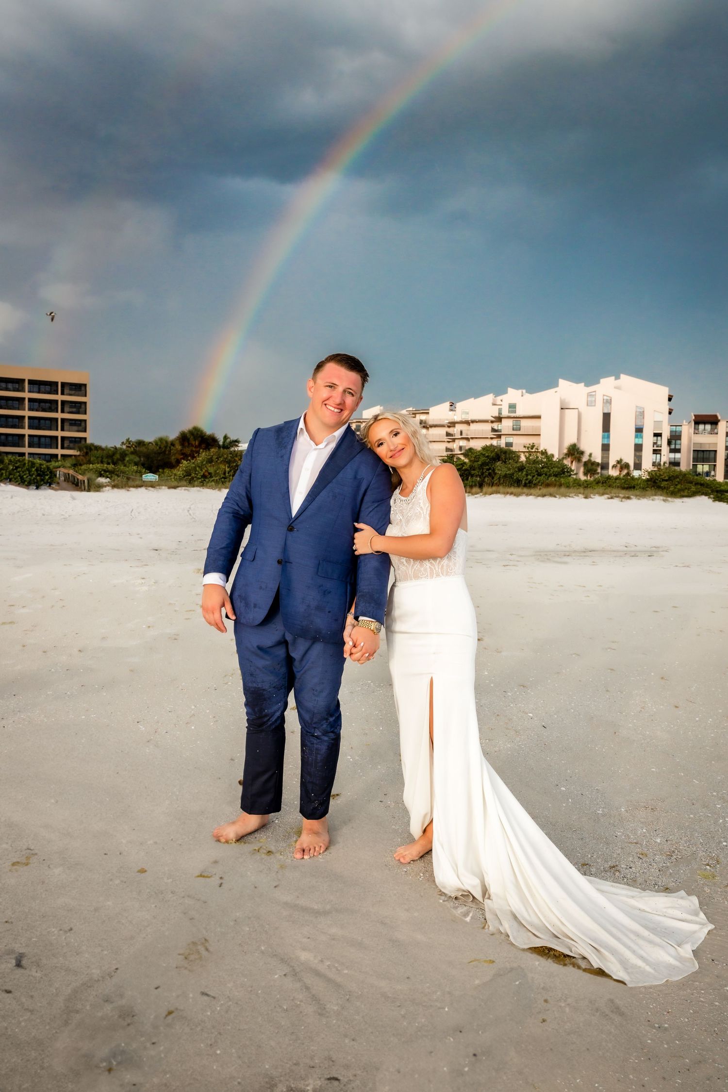 Wedding couple posing barefoot on white sand beach with a stunning rainbow arching overhead after the ceremony.