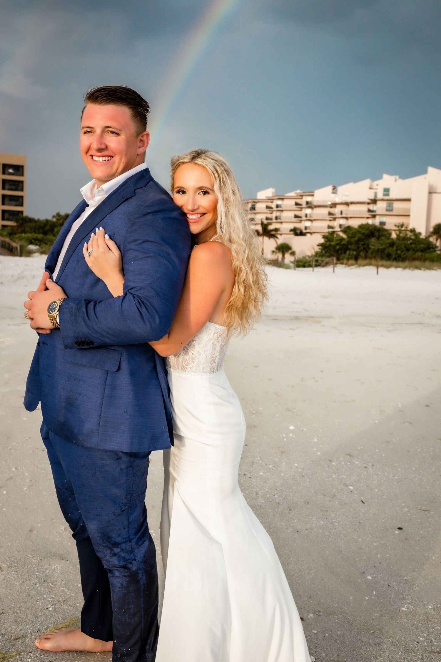 Smiling bride and groom standing back-to-back on a sandy beach with a rainbow visible in the background.