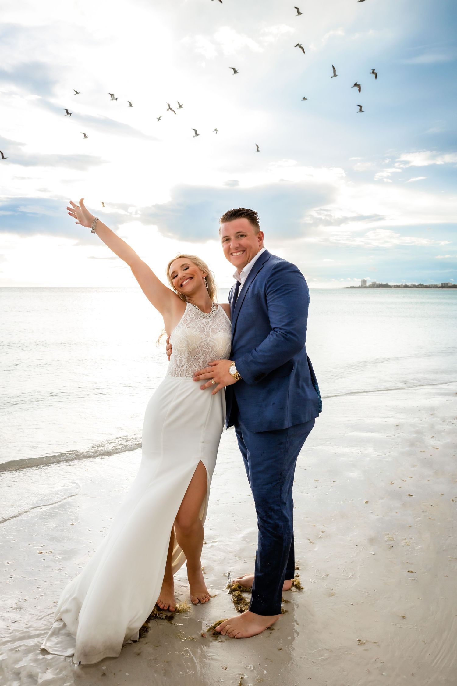 Joyful bride raising her arm with birds flying overhead as groom holds her on a beautiful beach wedding day.