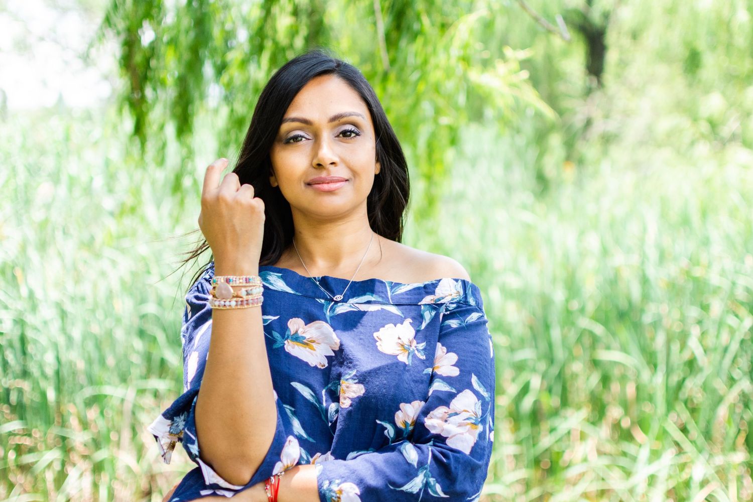 Woman in floral blue dress poses outdoors with willow trees in background.