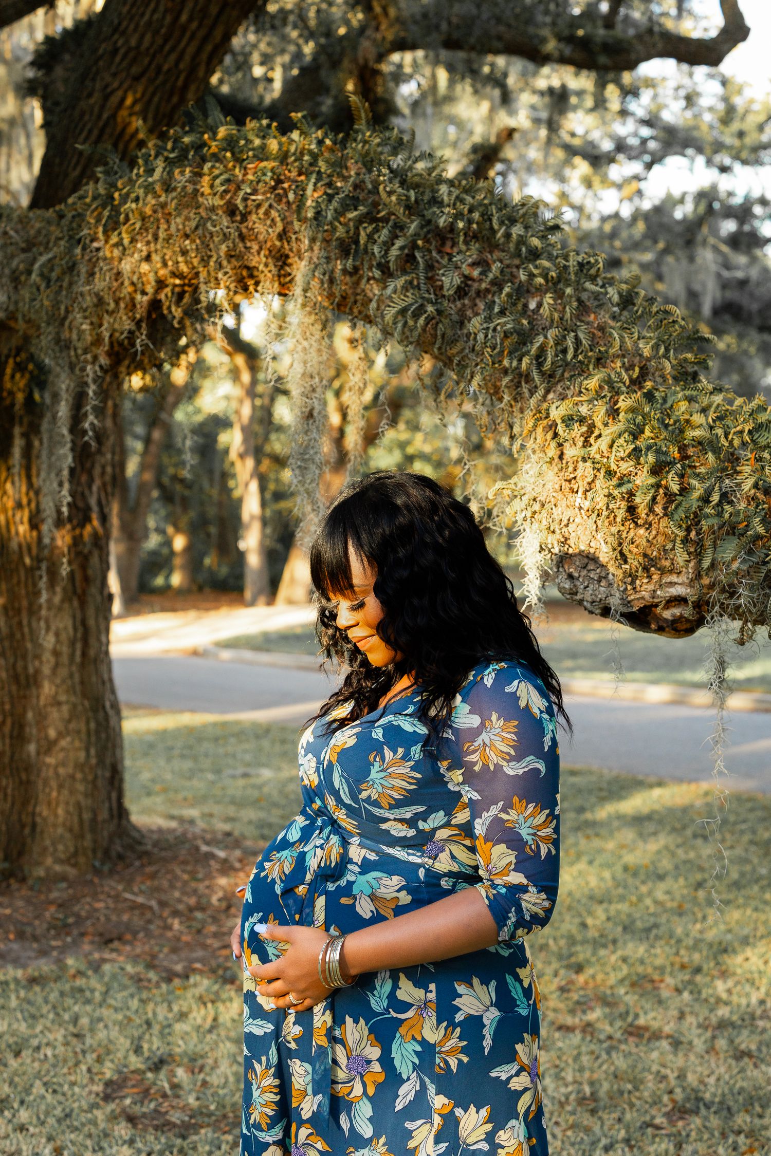 Pregnant woman in blue floral dress stands in golden afternoon light under mossy oak trees.