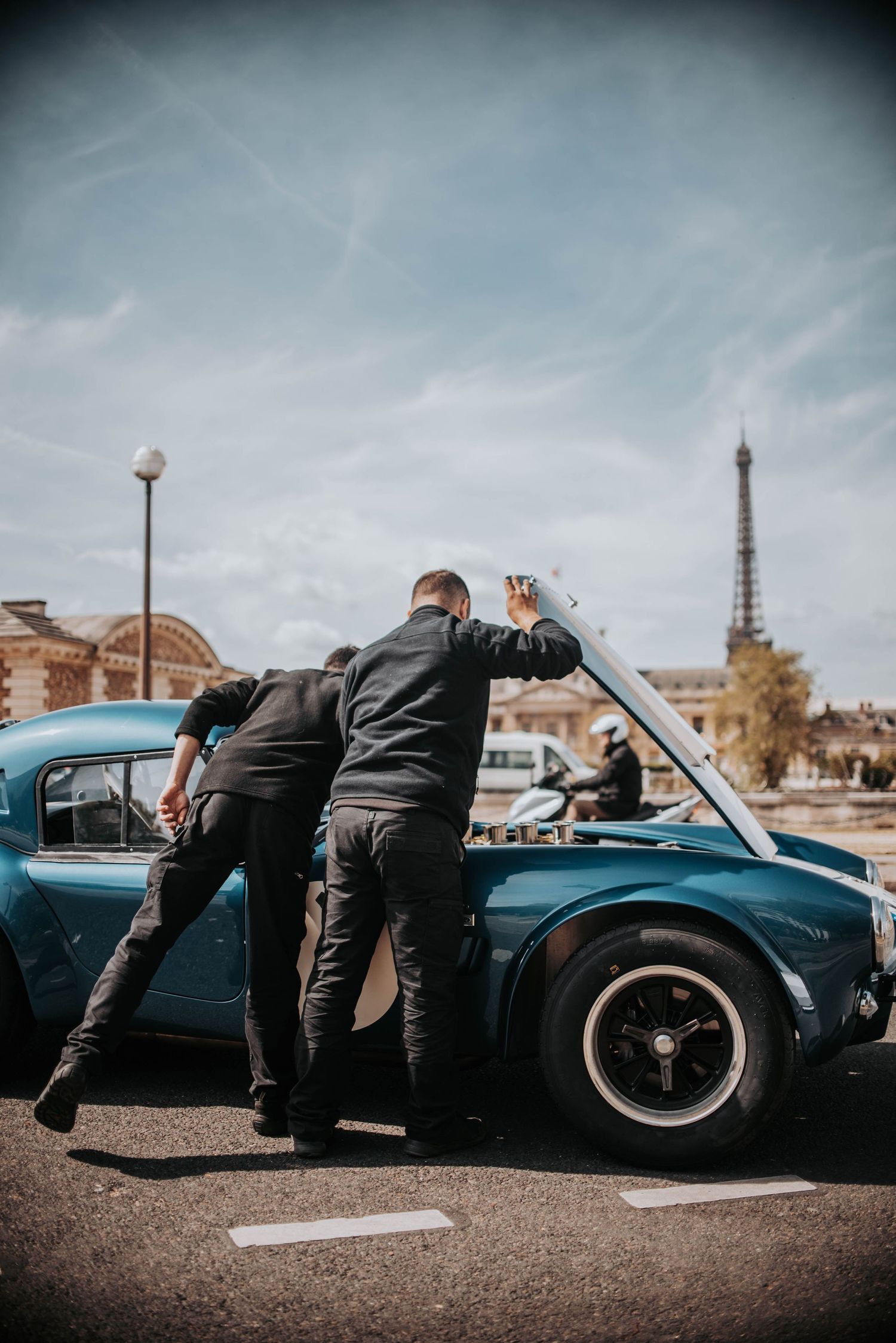People examining classic blue car with Eiffel Tower visible in background.