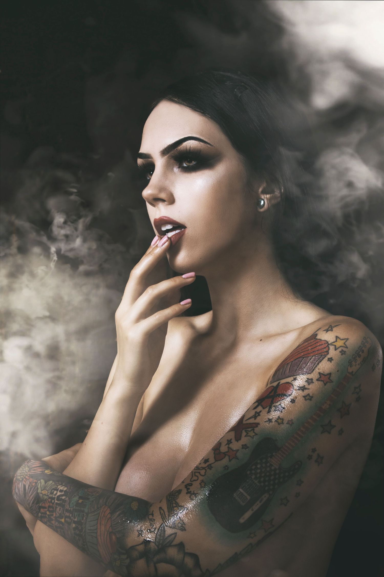 Artistic portrait with dark smoky atmosphere featuring dramatic makeup and tattoos in moody lighting.