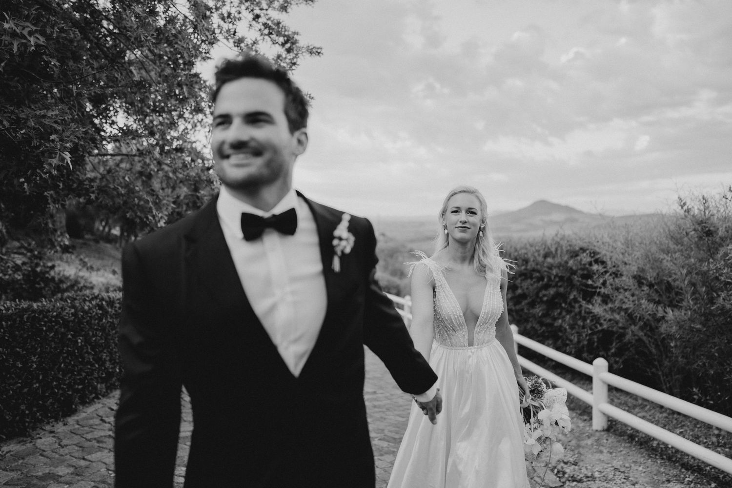 A wedding couple in black and white stands on a scenic overlook with mountain views and white fencing.