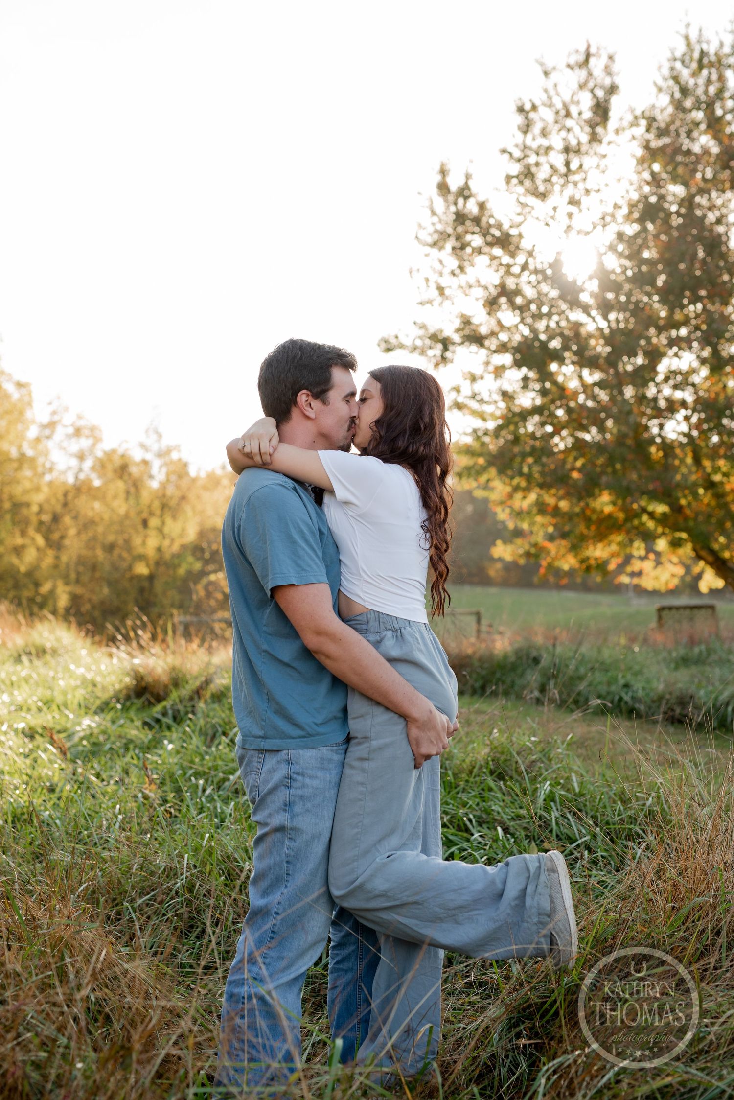 A couple embraces in a romantic pose on a grassy hillside during sunset.