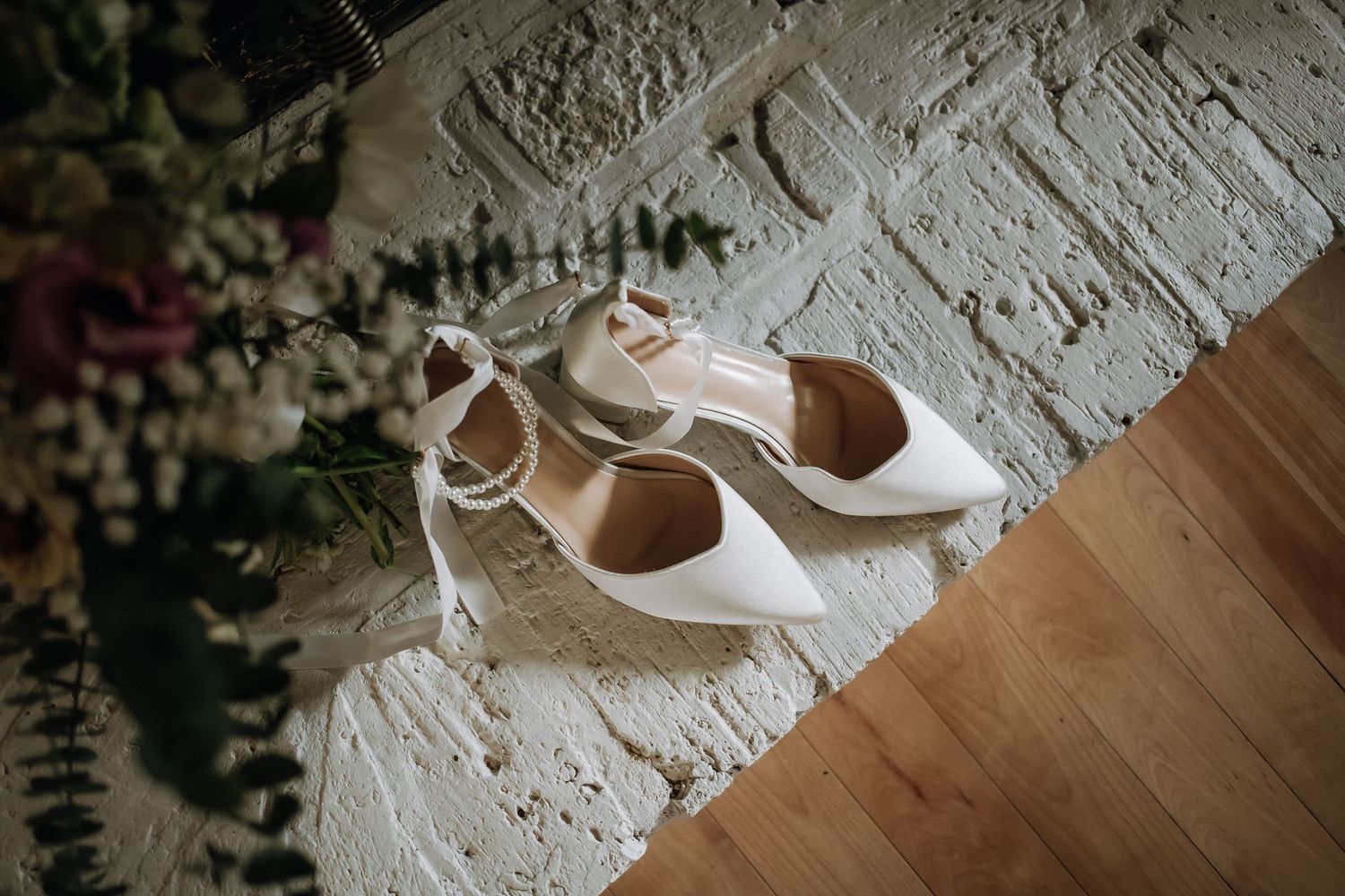 Elegant white bridal heels displayed on wooden floor next to white textured wall.
