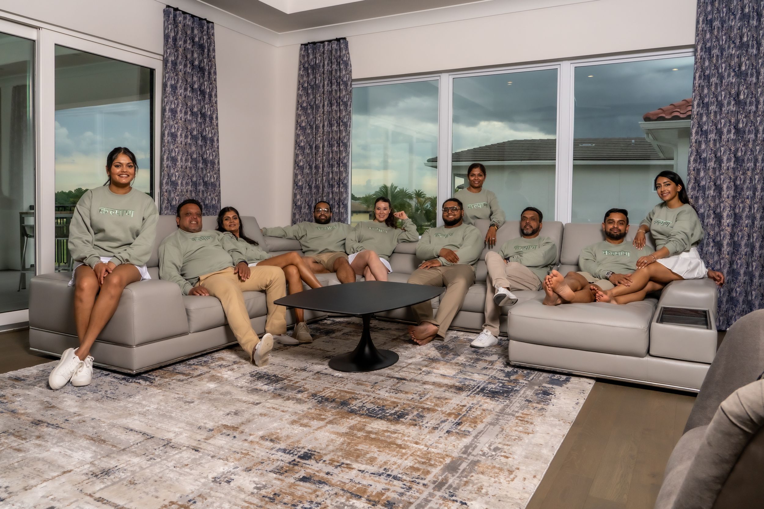 A group relaxes together on grey couches in a modern living room with blue curtains and patterned rug.