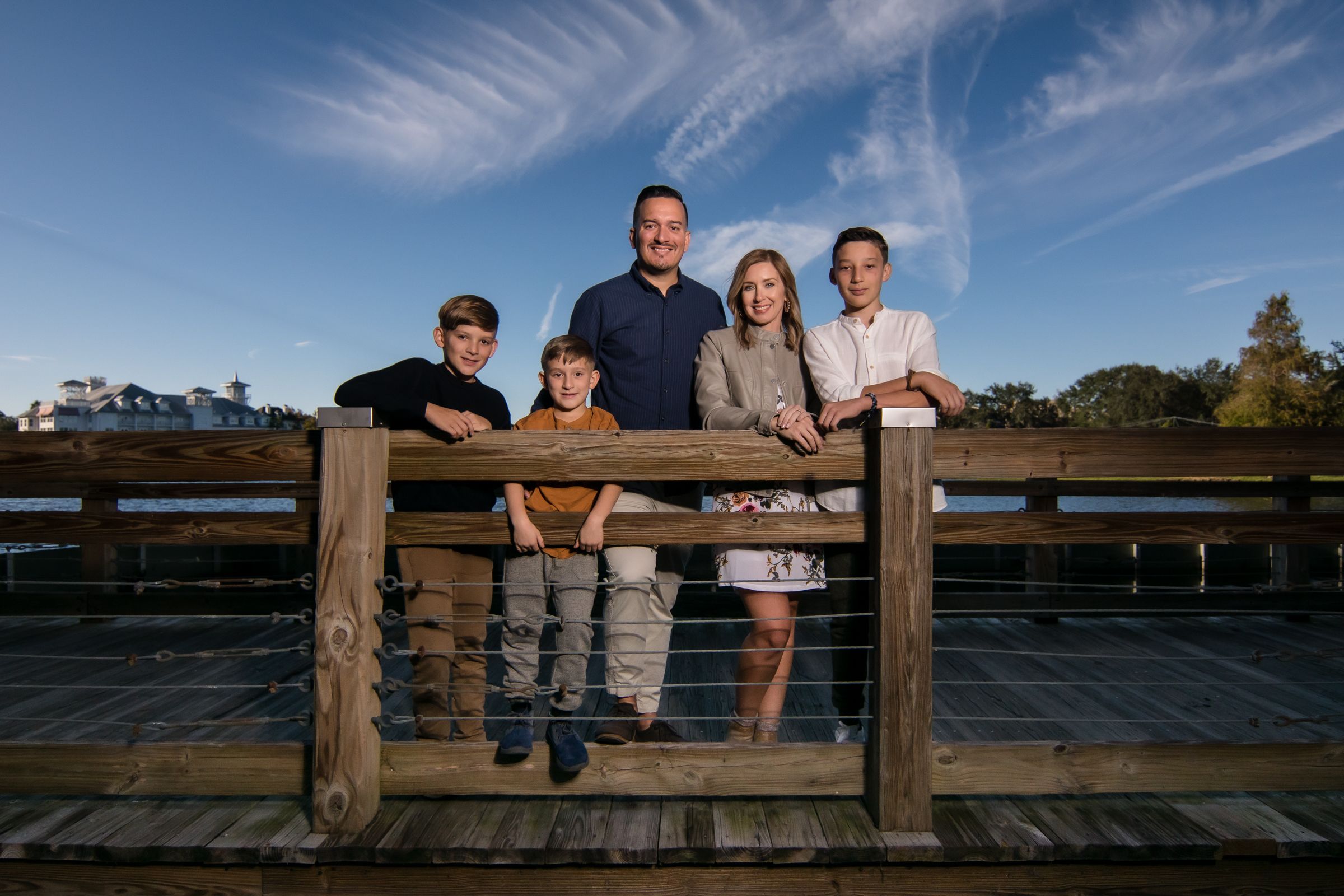 A family group poses on a wooden deck railing with blue sky and wispy clouds in the background.