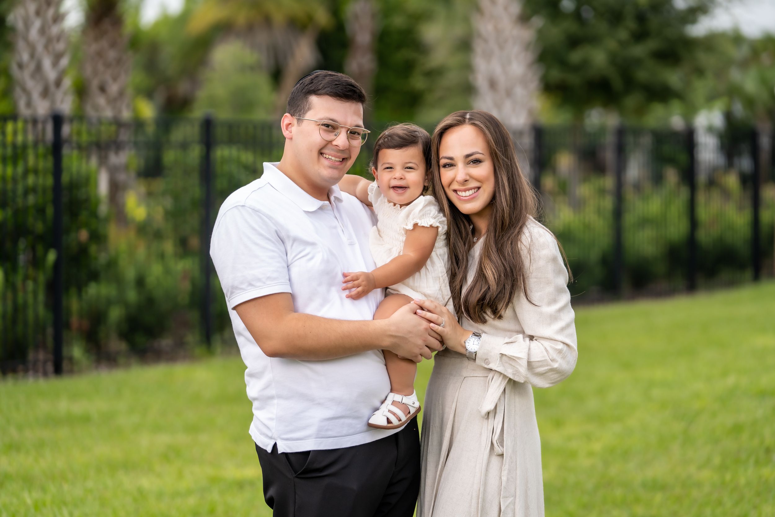 A young family poses together on a sunny day in front of a black fence and palm trees in a green garden.