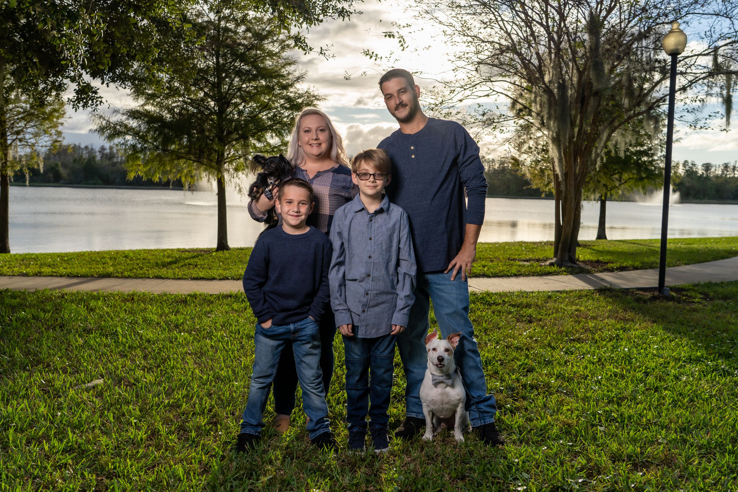 A family and their small grey dog pose together on grass with a lake in the background.