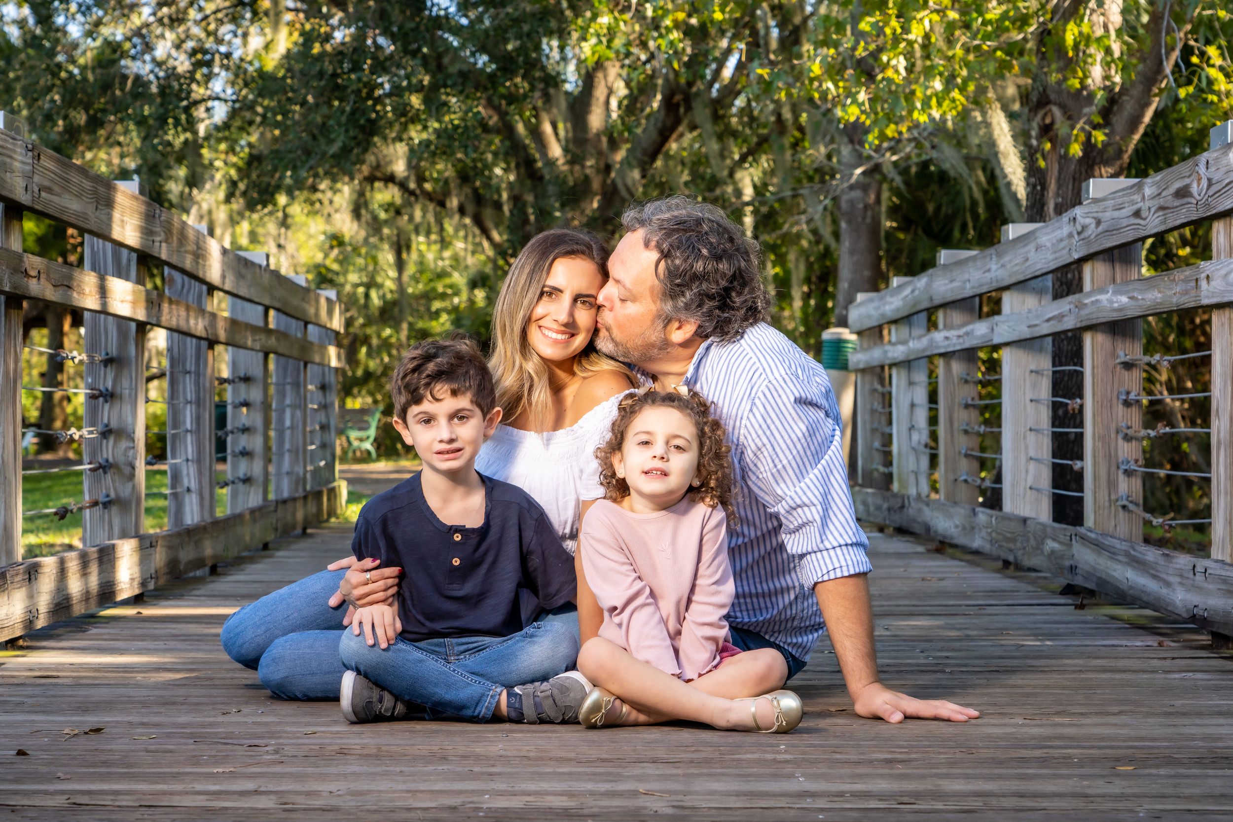 A family sits together on a wooden bridge surrounded by lush green trees in natural lighting.