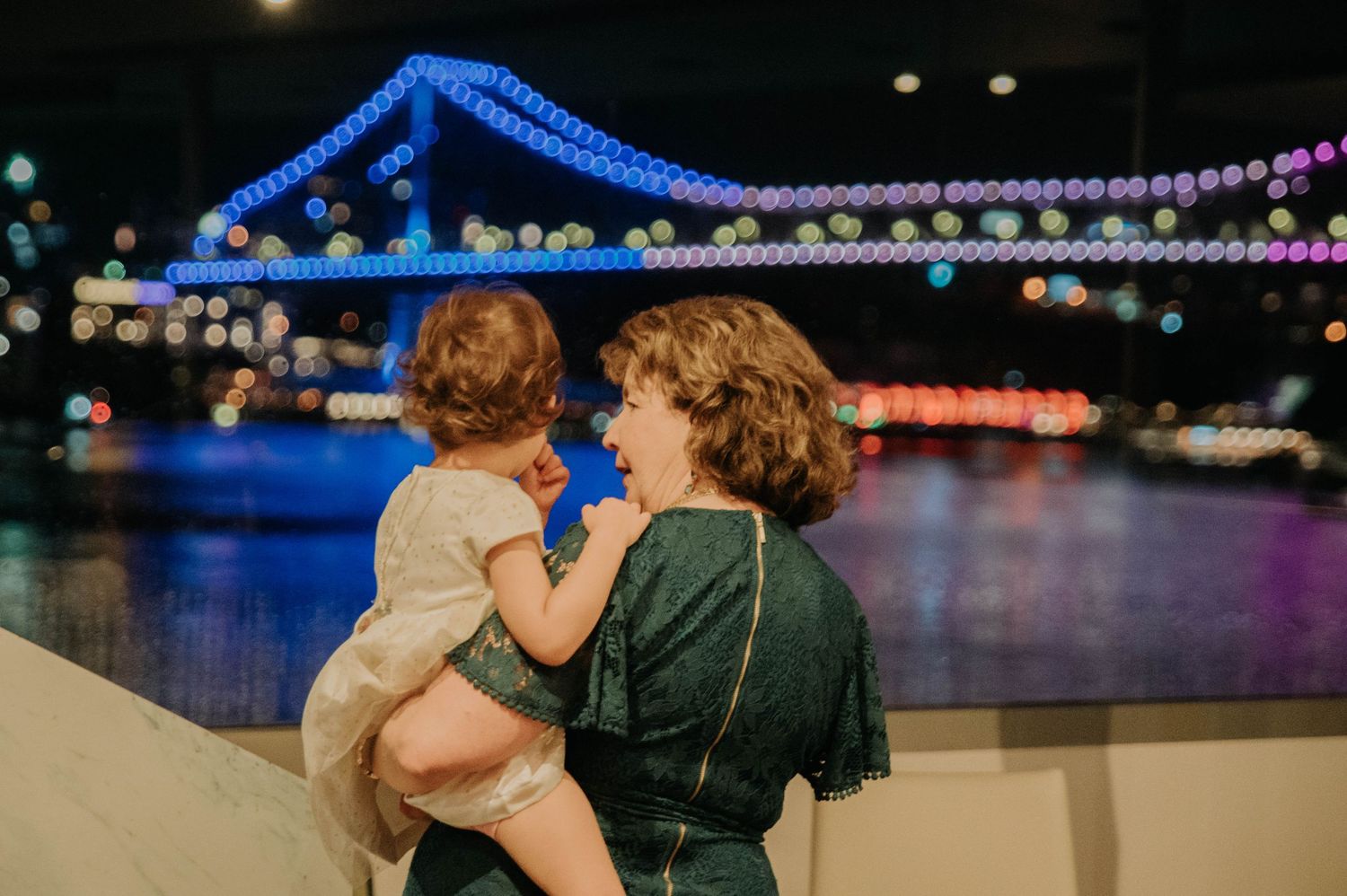 Mother in green dress holds child while enjoying illuminated nighttime bridge view.
