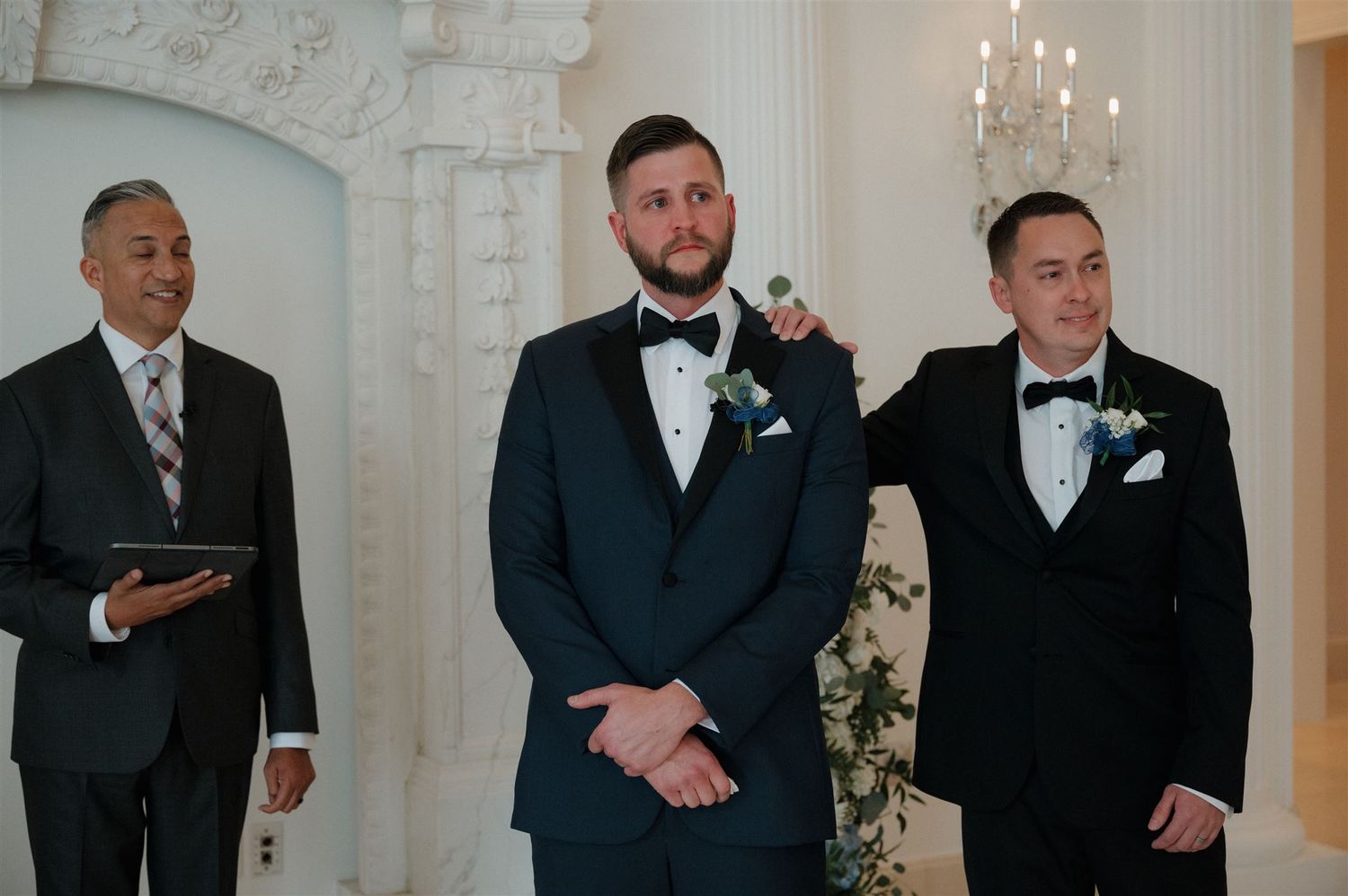 Three people wearing black tuxedos and bow ties stand at front of elegant white wedding chapel with chandelier.