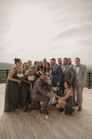 Wedding party poses playfully for group photo on wooden deck with mountains in background.