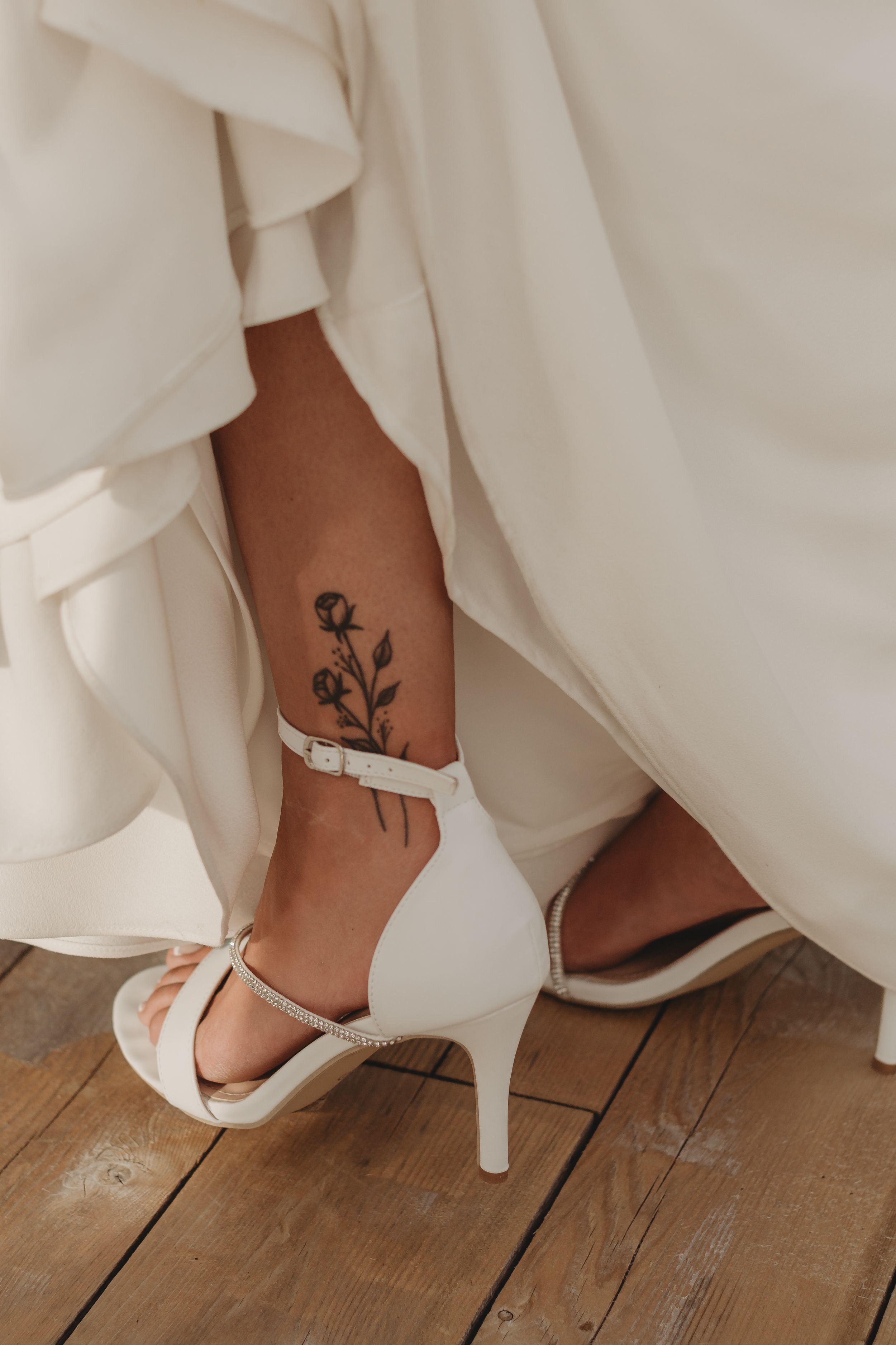 Close-up of bridal shoes and floral ankle tattoo against wooden deck flooring.