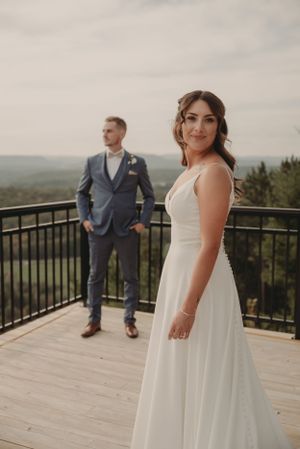 Wedding couple stands on wooden deck with scenic mountain backdrop during outdoor photo session.