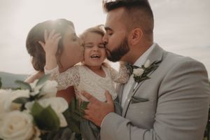 Sweet family moment at wedding with couple kissing small child in lace dress.