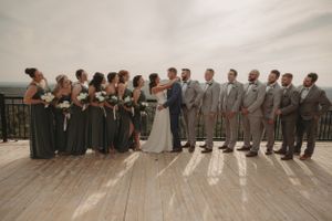 Wedding party lined up on outdoor deck wearing sage green dresses and gray suits against mountain backdrop.